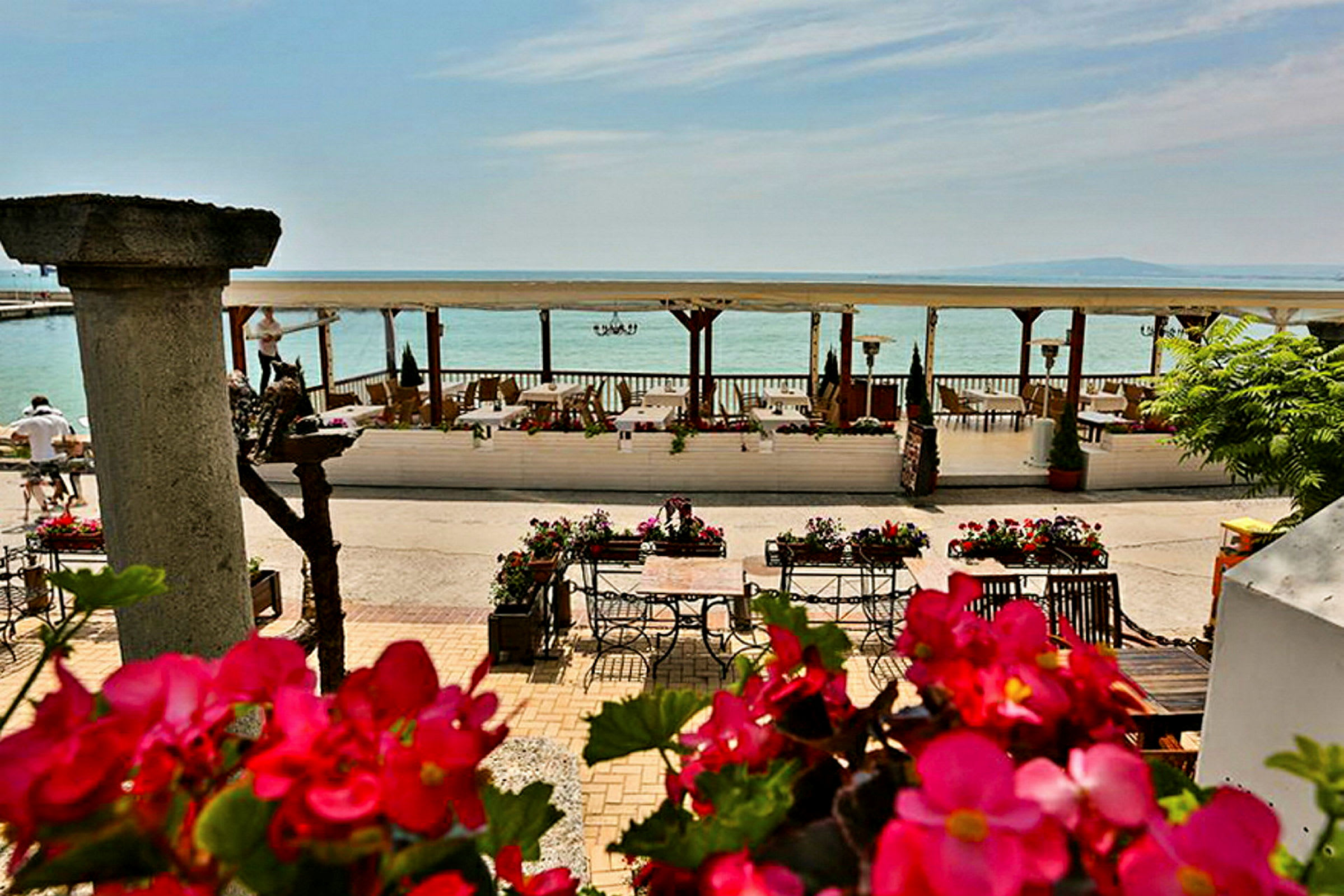 En strandnær terrasse med bord og stoler omgitt av blomster og utsikt over havet. (AI-generert tekst)