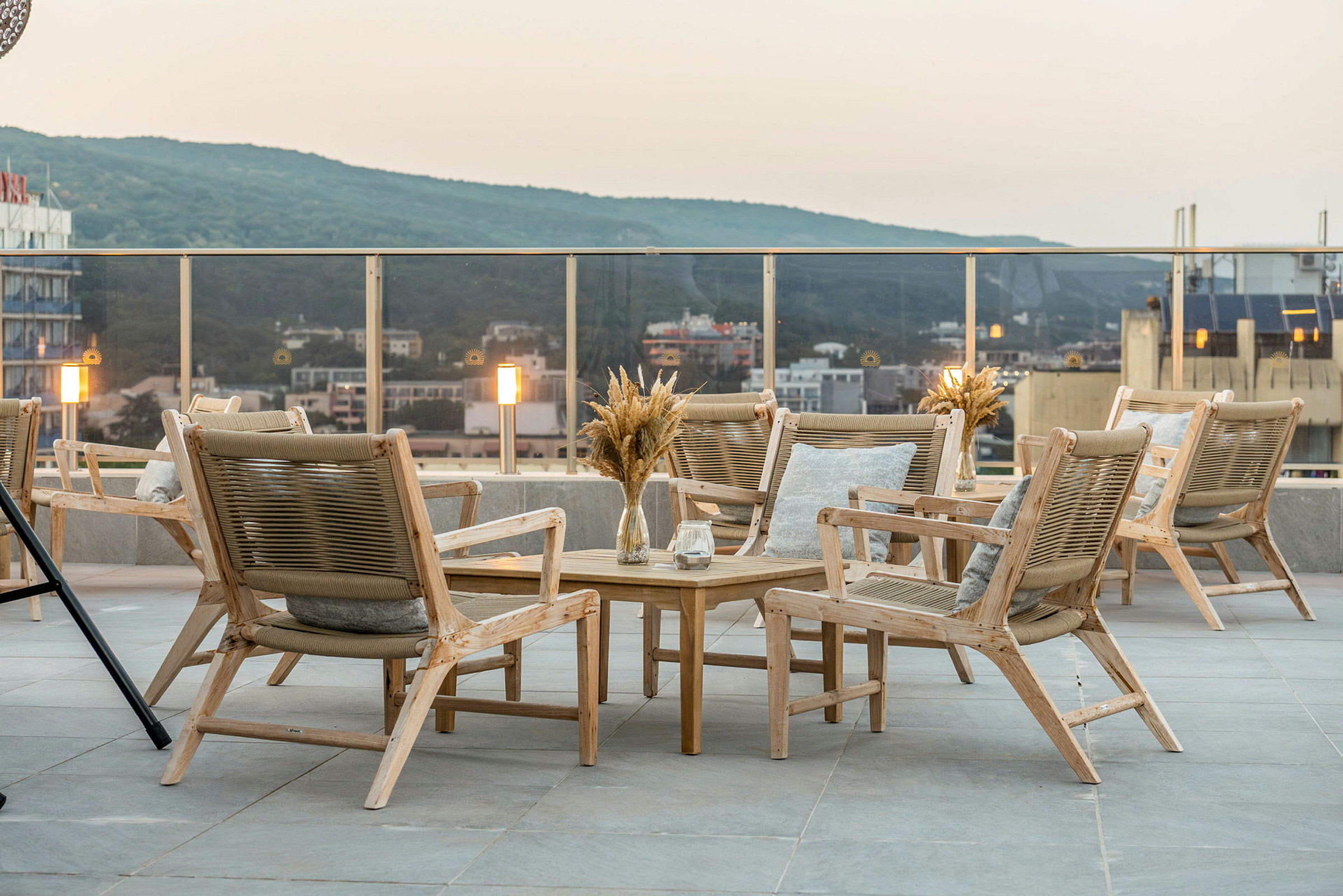 Terrasse med en gruppe tremøbler, inkludert stoler og bord. Utsikt over byen og grønne åser i bakgrunnen. (AI-generert tekst)