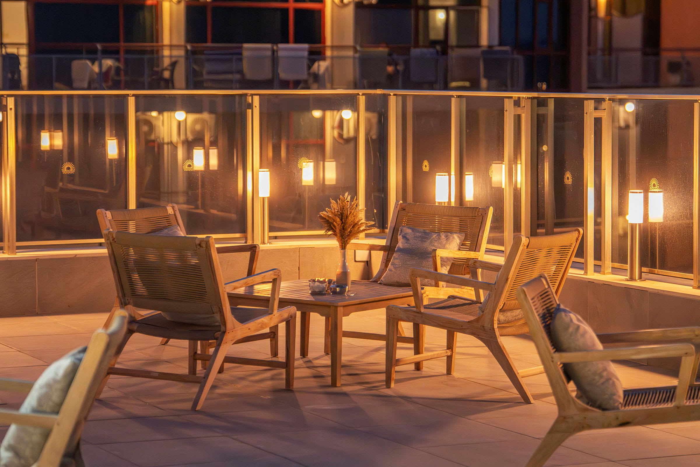 Fire stoler i tre med puter er plassert rundt et lite bord på en terrasse med glassrekkverk og belysning. (AI-generert tekst)
