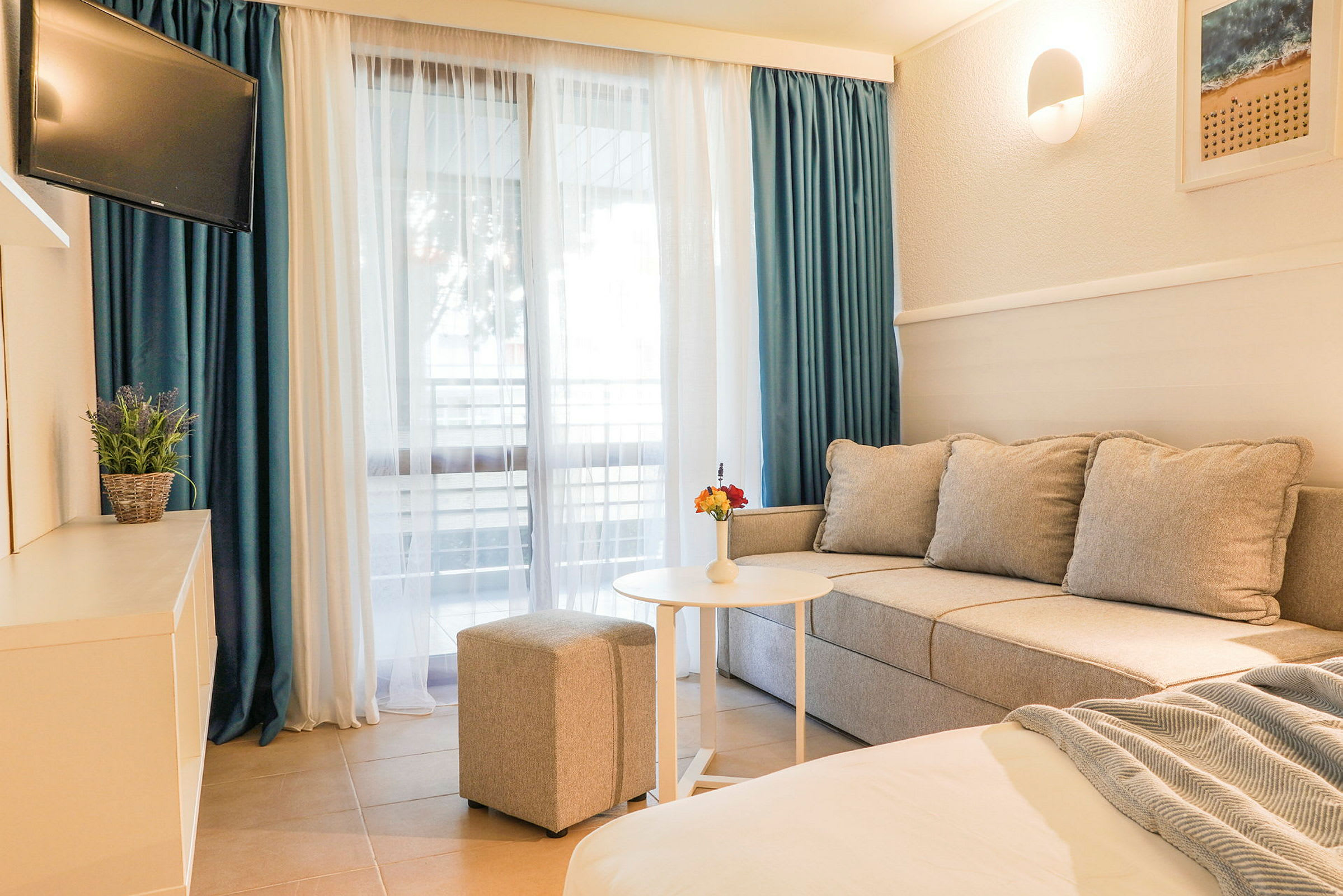 Et hotellrom med sofa, bord, puff og TV, samt et stort vindu med gardiner. (AI-generert tekst)