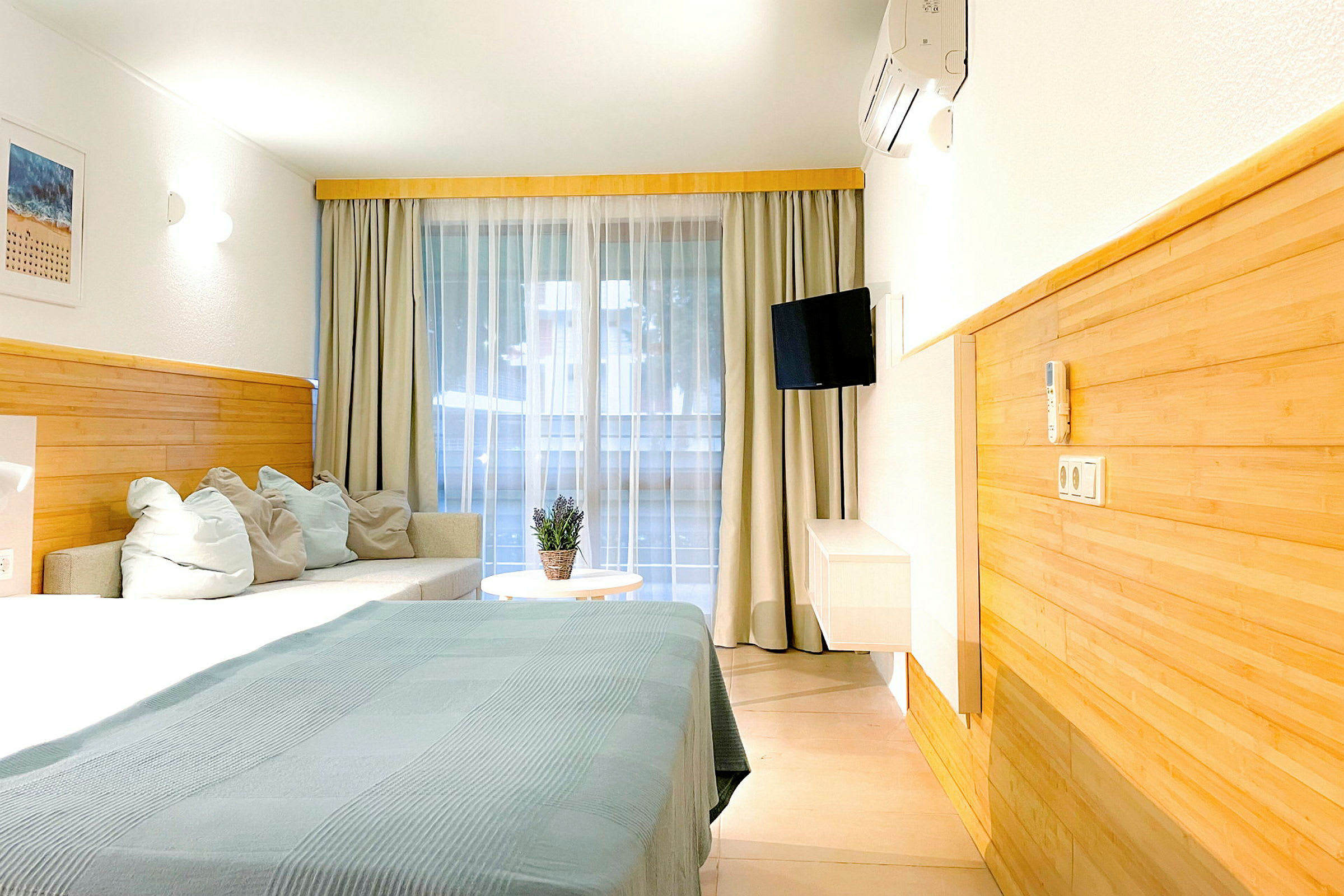 Et hotellrom med seng, sofa og bord. Gardiner henger foran et stort vindusområde. (AI-generert tekst)