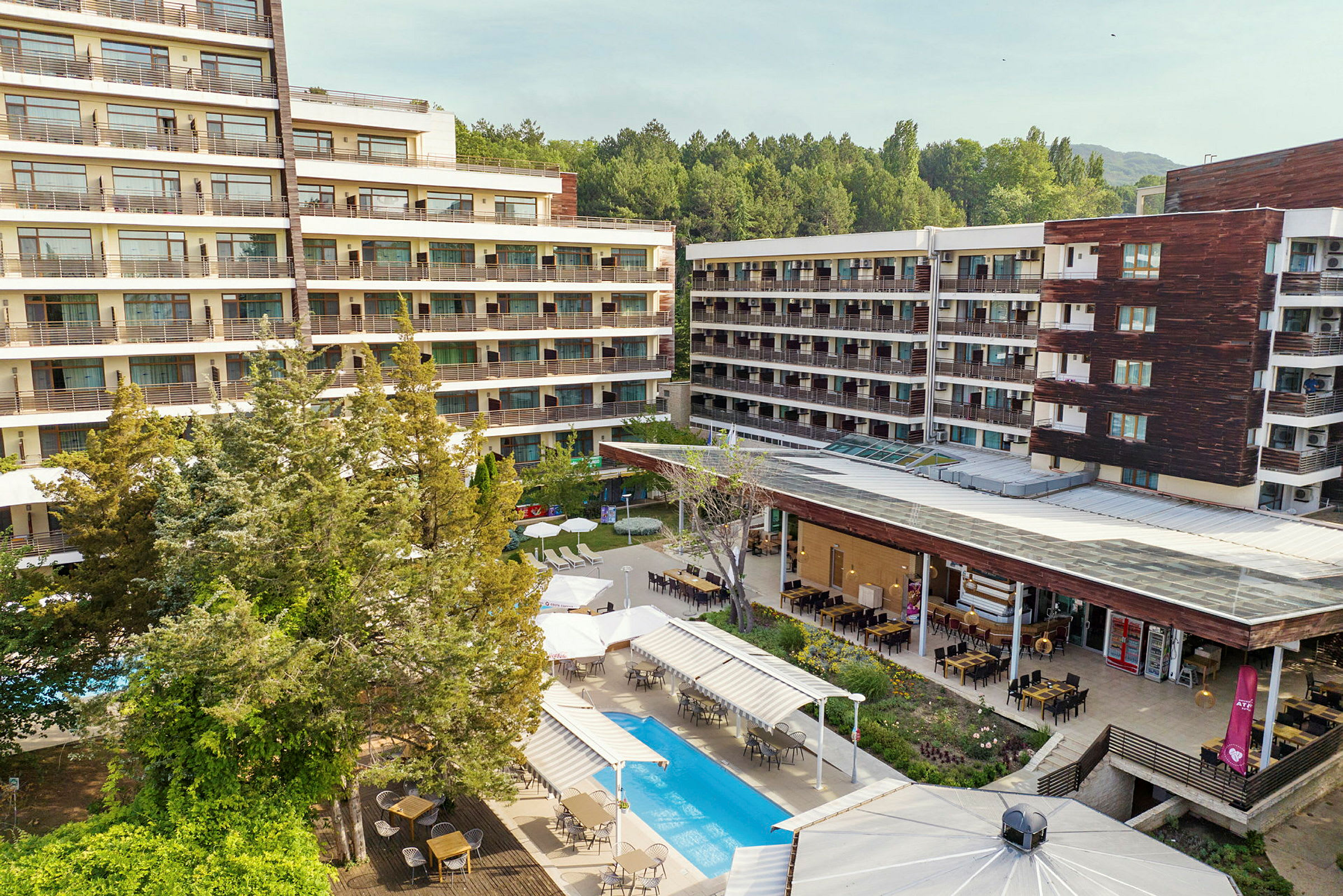 Hotellbygninger omgitt av grøntområder. Basseng og utendørs spiseområde vises i forgrunnen. (AI-generert tekst)