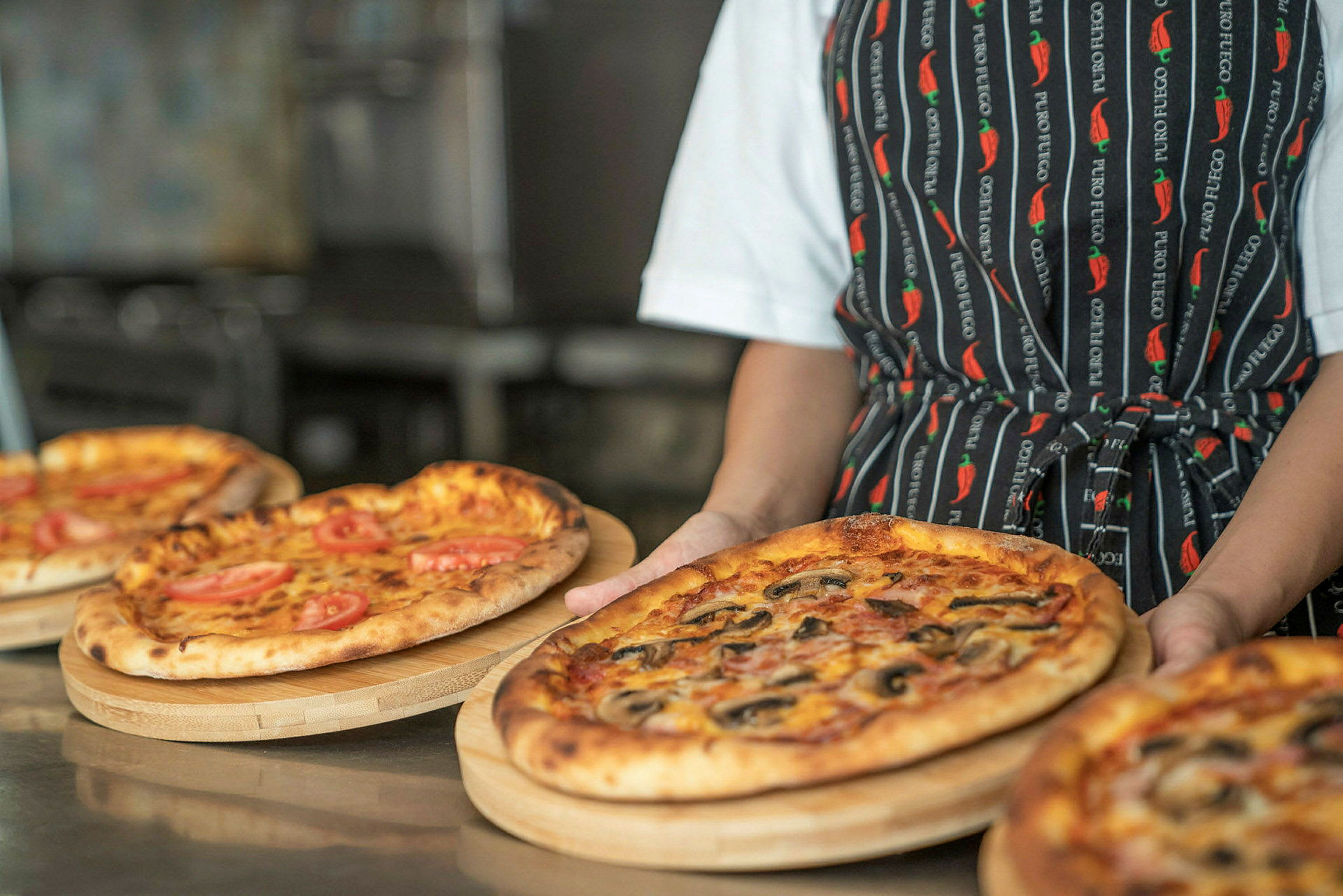 En person holder et trebrett med en pizza, omgitt av andre pizzaer. (AI-generert tekst)
