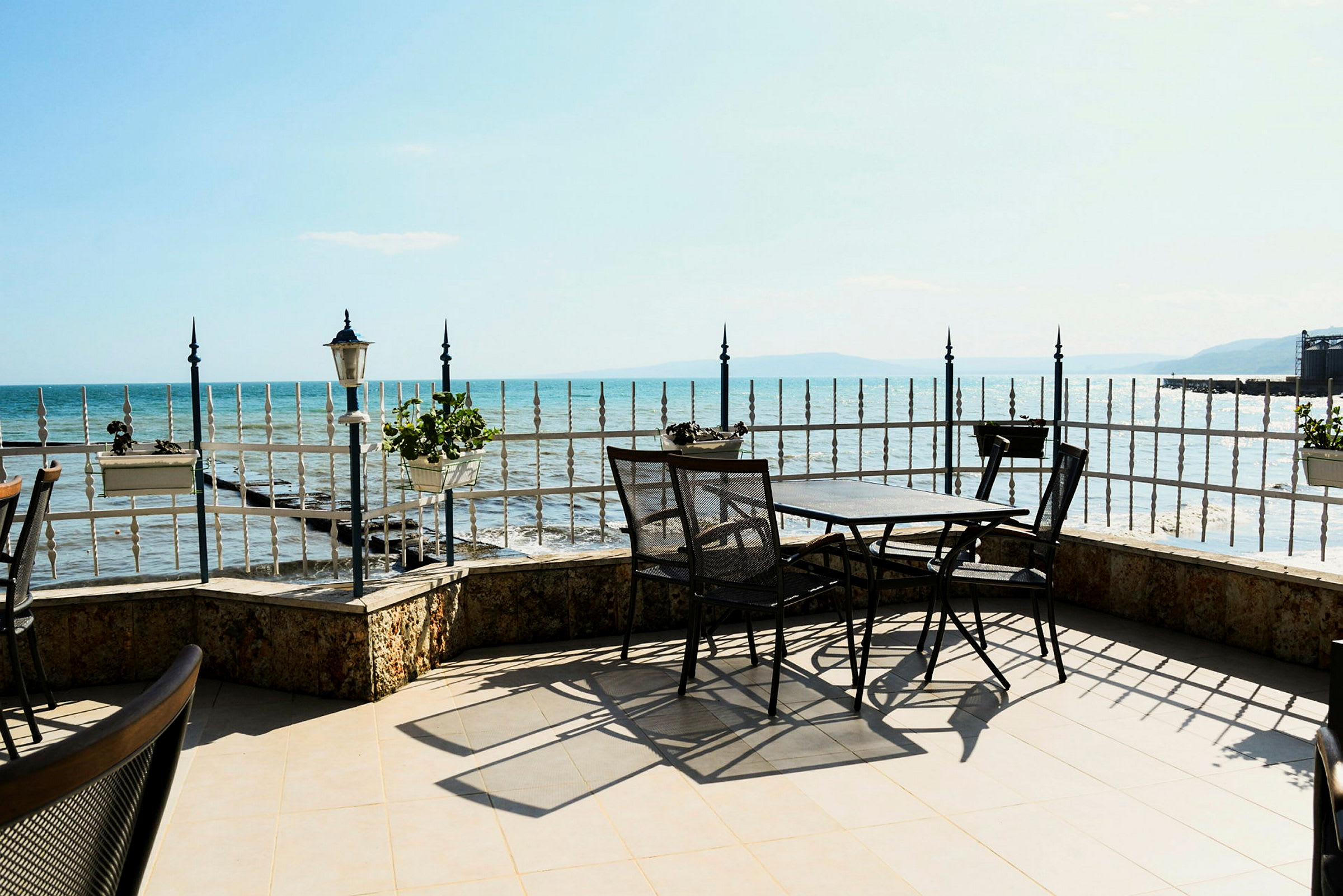 Terrasse med bord og stoler ved et jerngjerde, med utsikt over havet under klart vær. (AI-generert tekst)