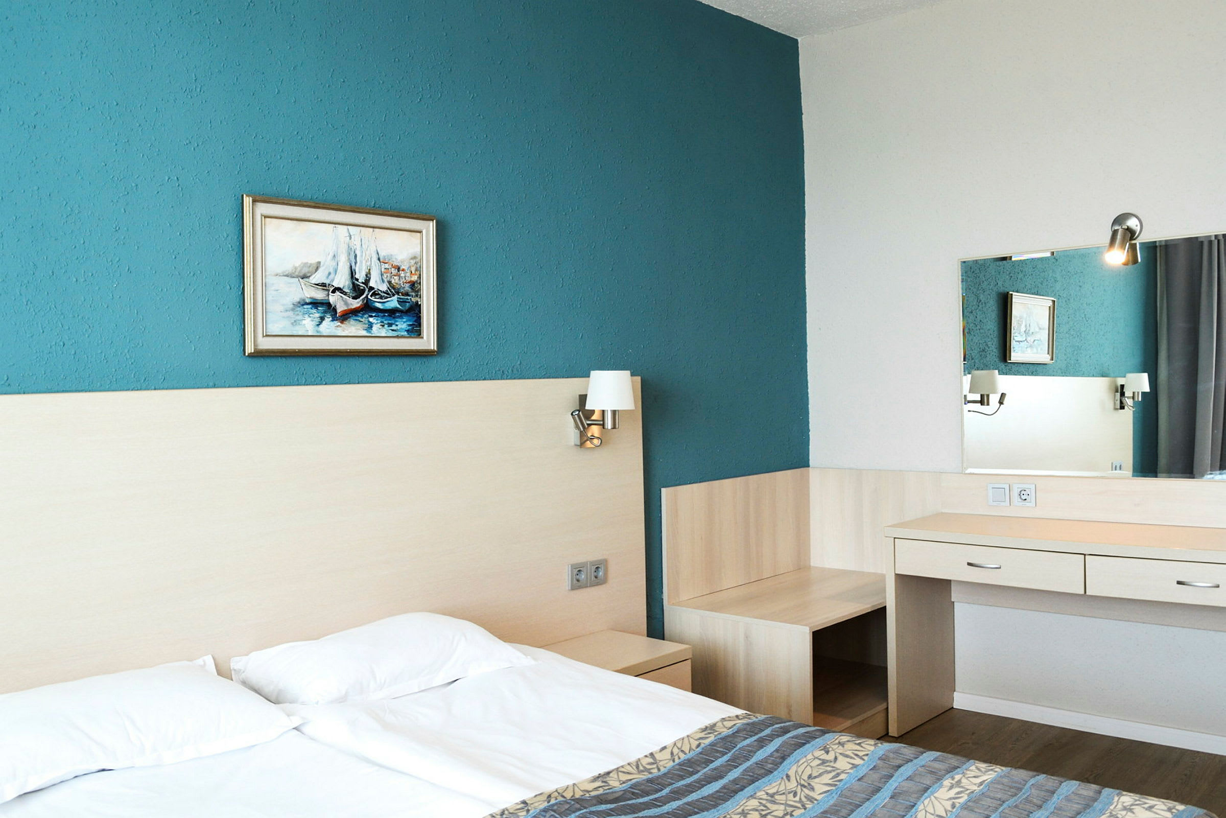 Hotellrom med dobbeltseng, blå vegg, maleri, senge lamper og skrivebord med speil. (AI-generert tekst)