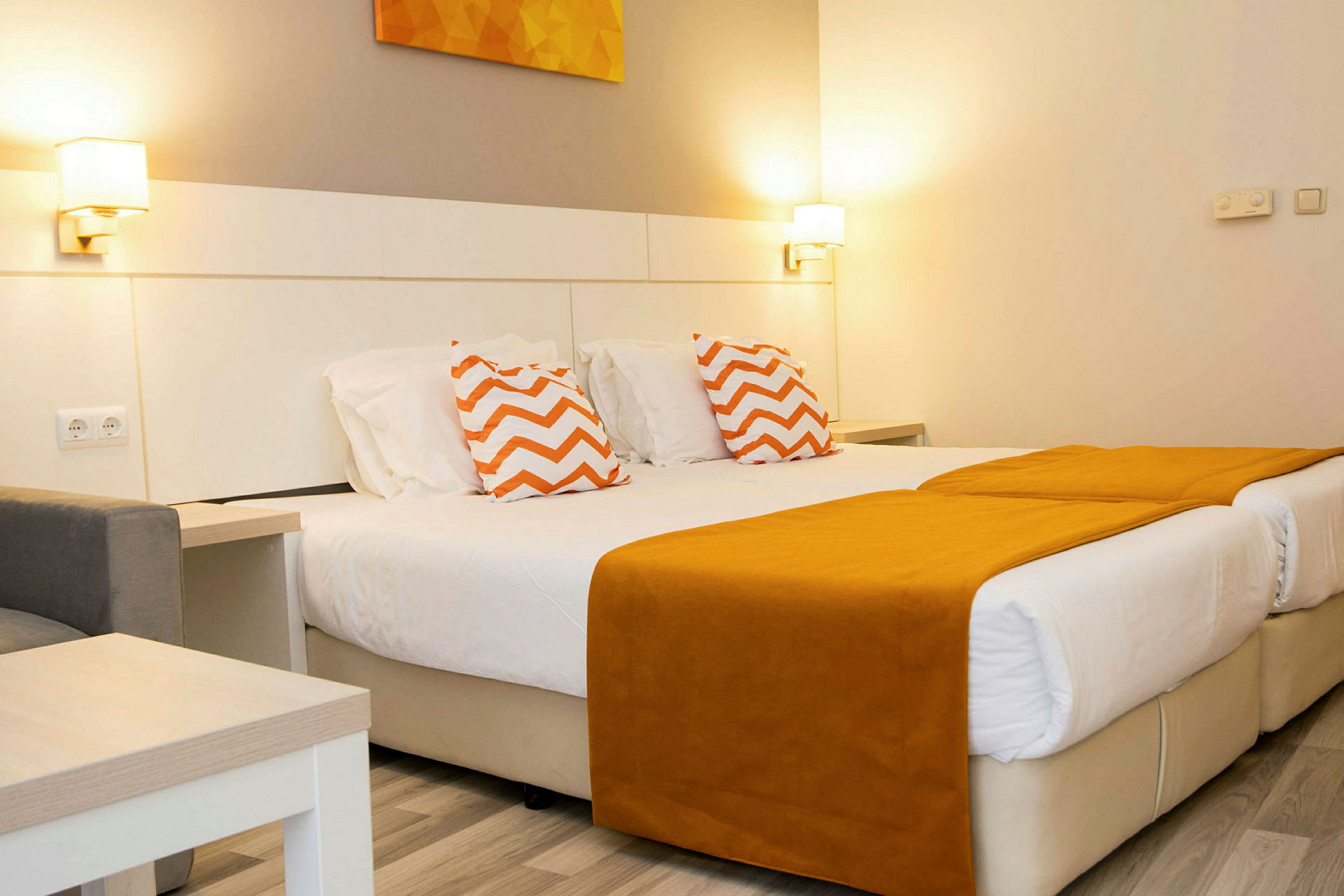 Et hotellrom med to enkeltsenger med oransje sengetepper og pynteputer med sikksakkmønster. (AI-generert tekst)