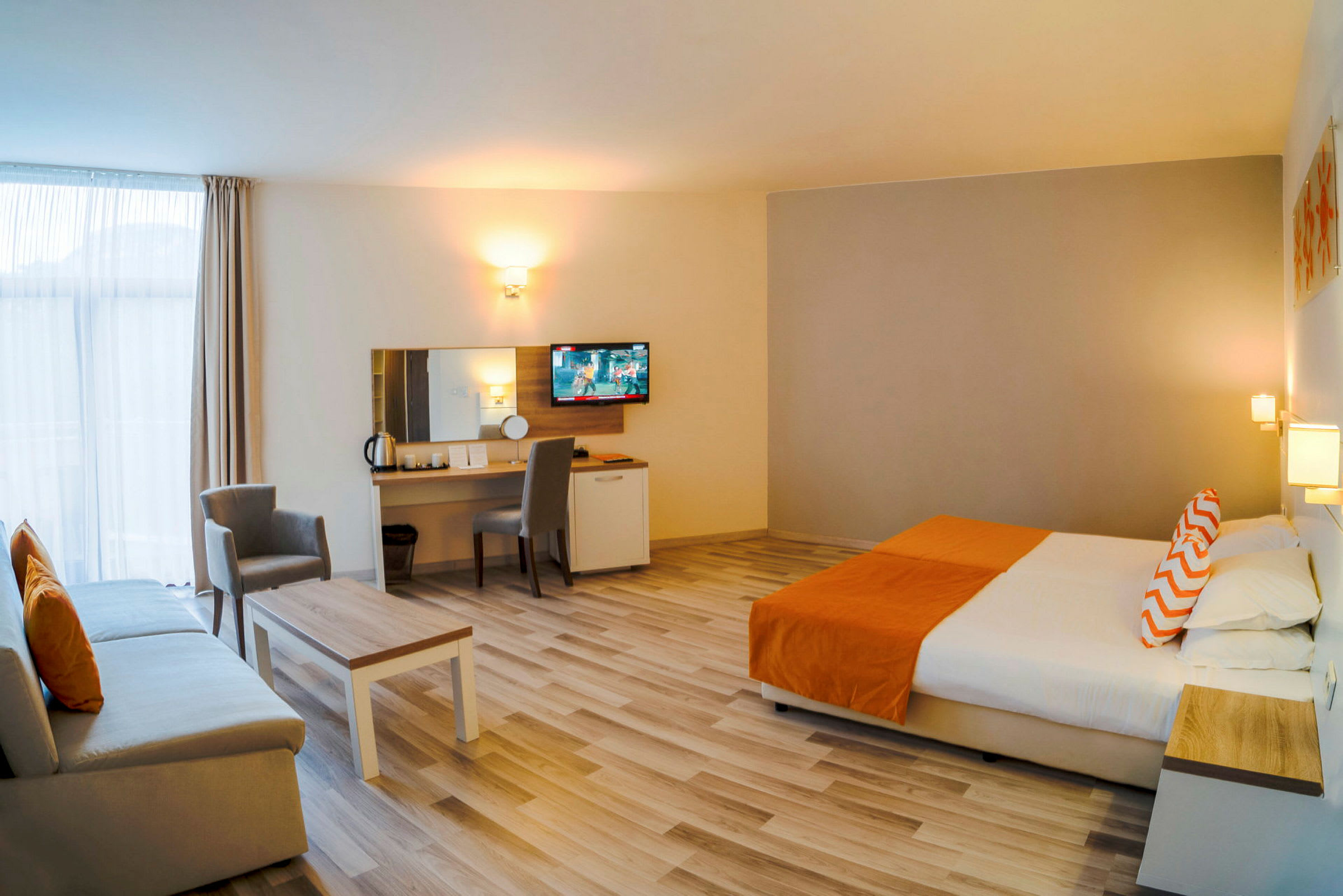 Bilde av et romslig hotellrom med en stor seng, sofa, bord og arbeidsområde, innredet i nøytrale og oransje toner. (AI-generert tekst)