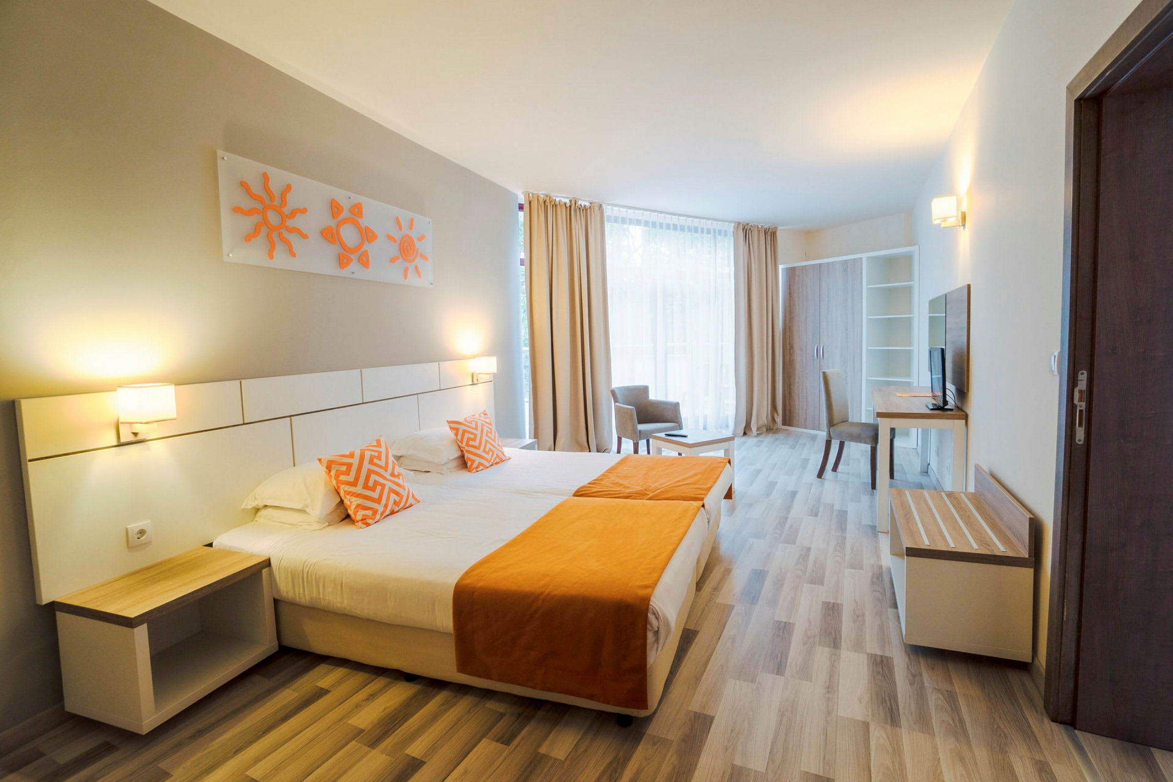 Et hotellrom med en dobbeltseng som har oransje detaljer, to senge lamper og moderne interiør. En bred vindusflate med lyse gardiner ses i bakgrunnen. (AI-generert tekst)