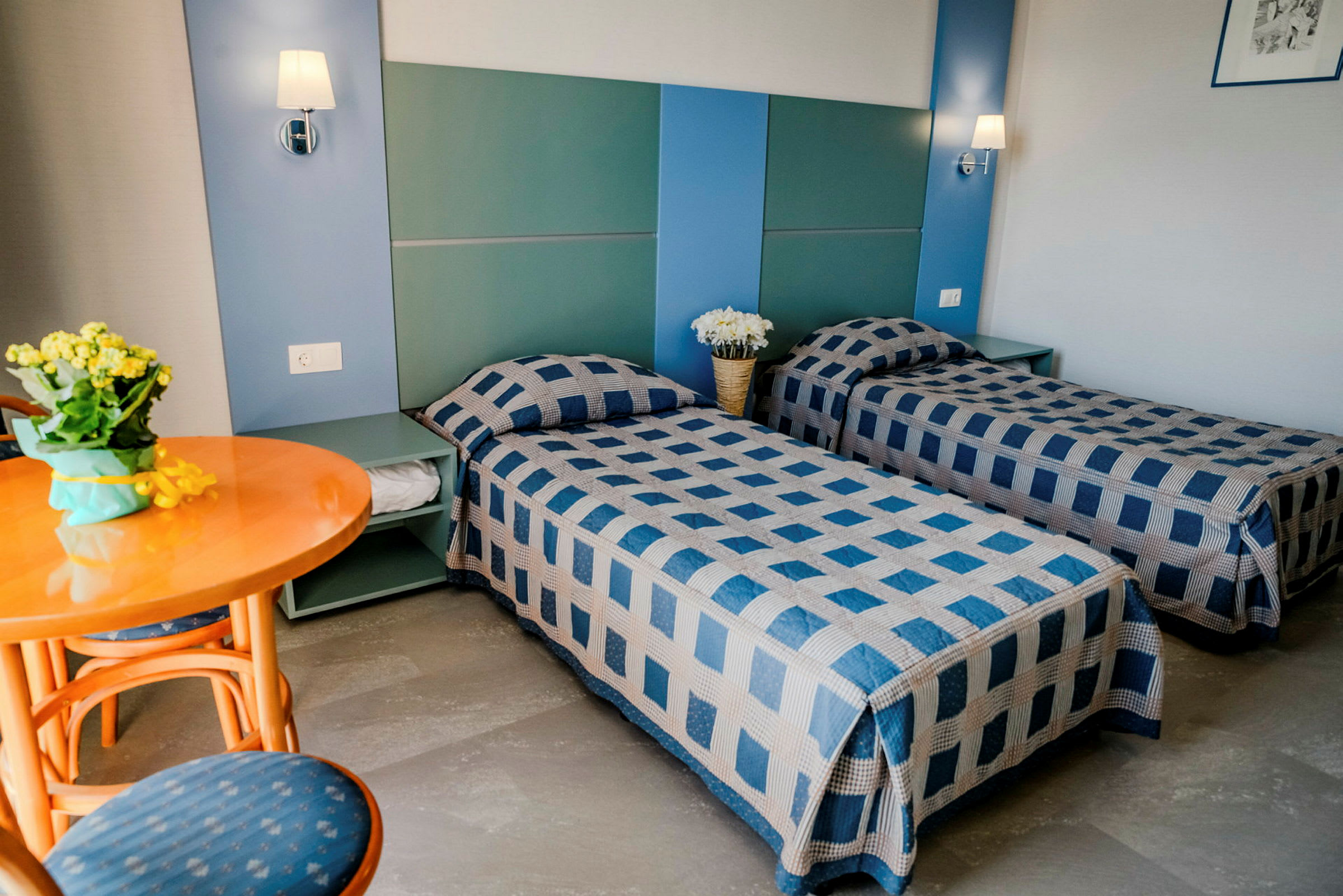 Et hotellrom med to enkeltsenger med blåmønstrede sengetepper, et rundt trebord med stoler samt dekorasjoner på nattbordet. (AI-generert tekst)