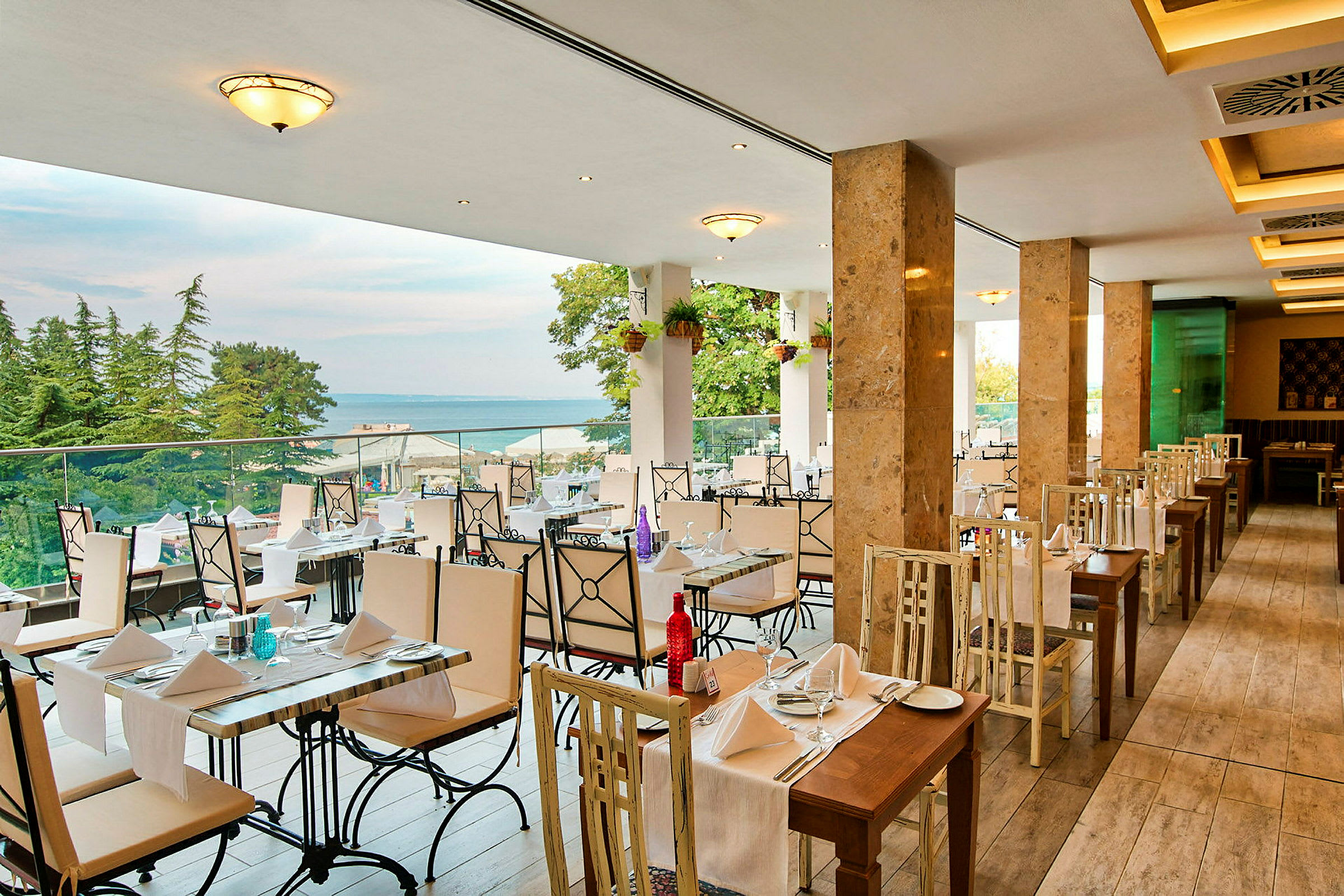 En restaurant med bord og stoler i lyse farger, utsikt over havet og omgitt av trær. (AI-generert tekst)