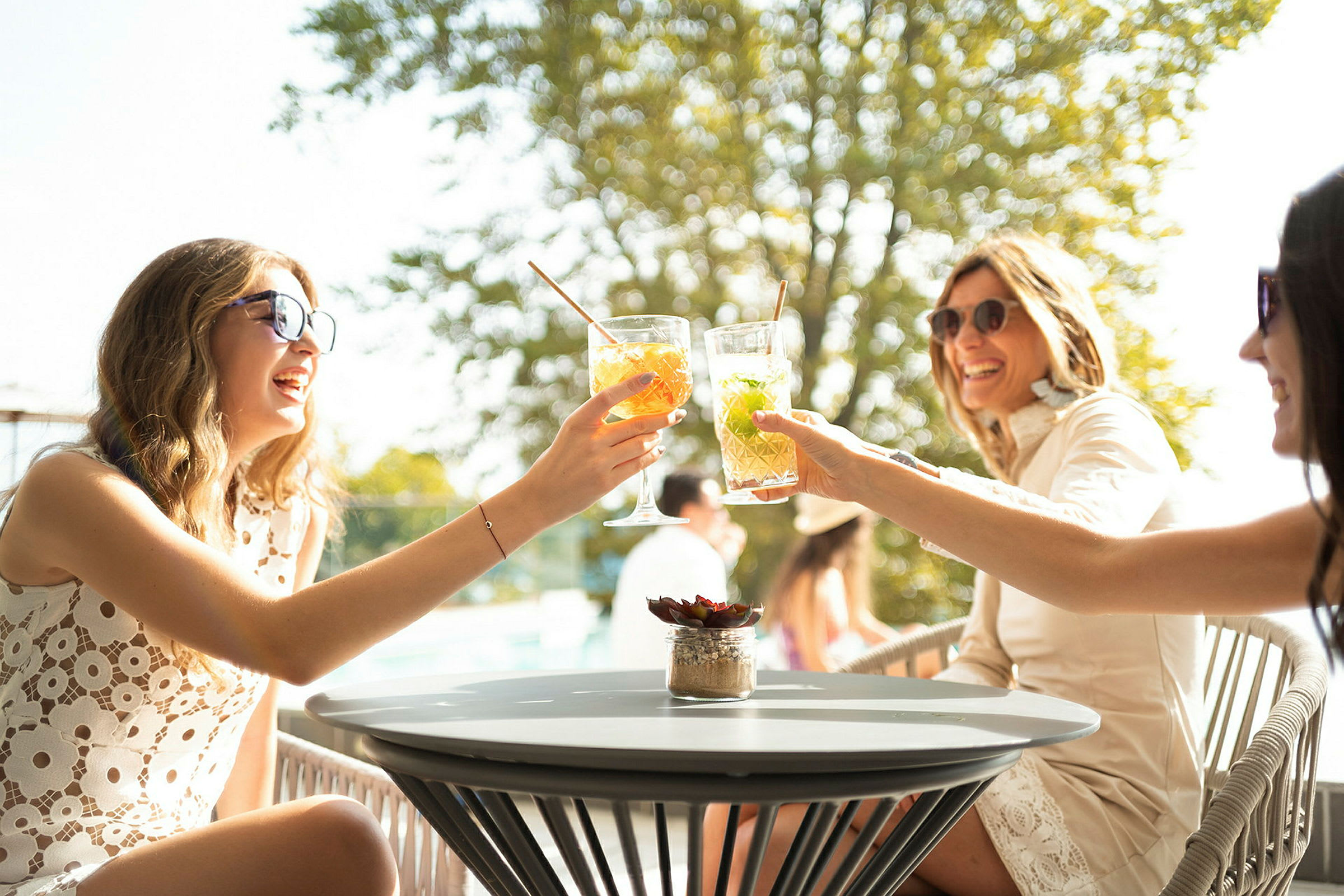 Personer som skåler med drinker ved et bord utendørs med trær i bakgrunnen. (AI-generert tekst)