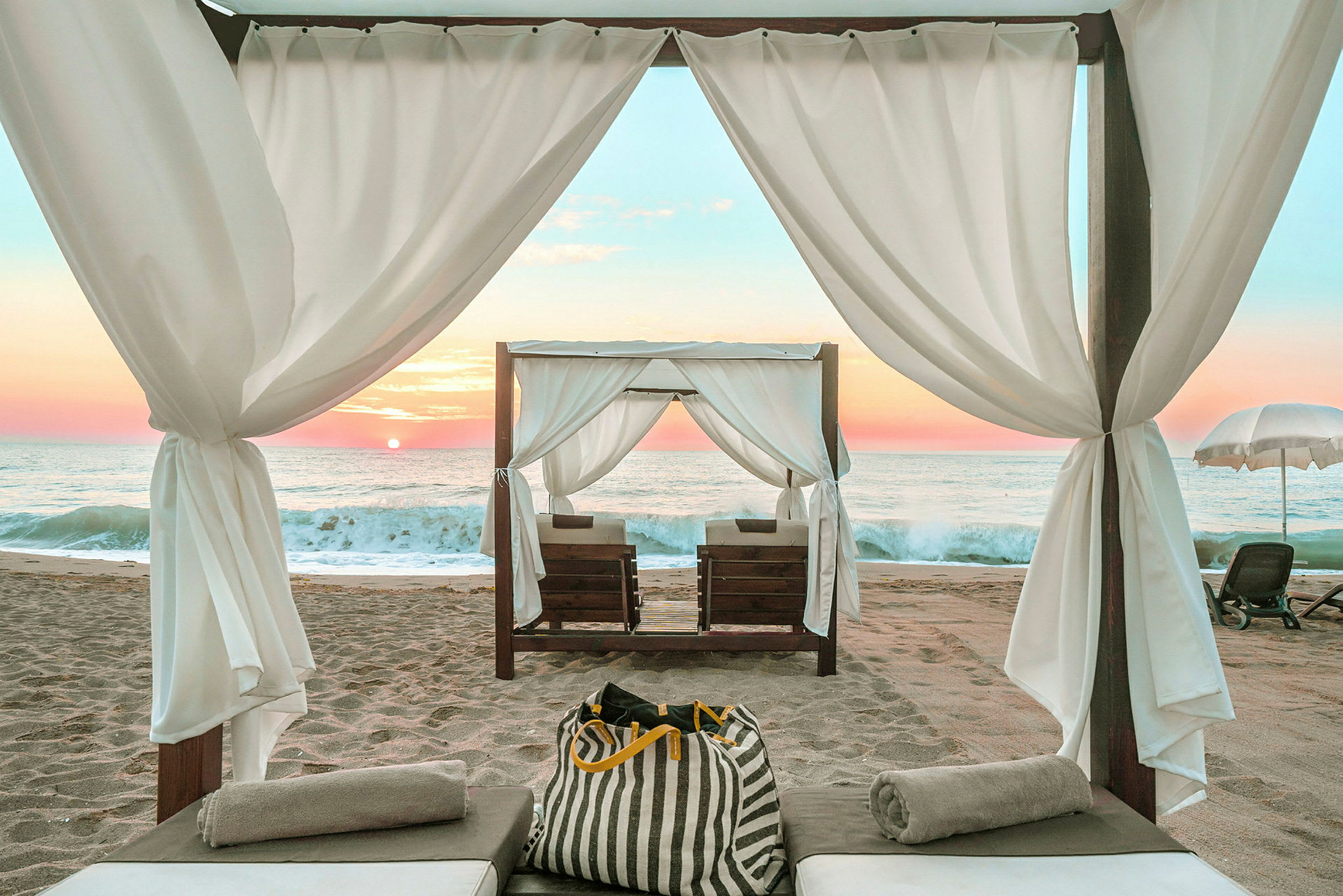 En oppredd solseng under en skyggefull cabana på en sandstrand ved havet ved solnedgang. (AI-generert tekst)