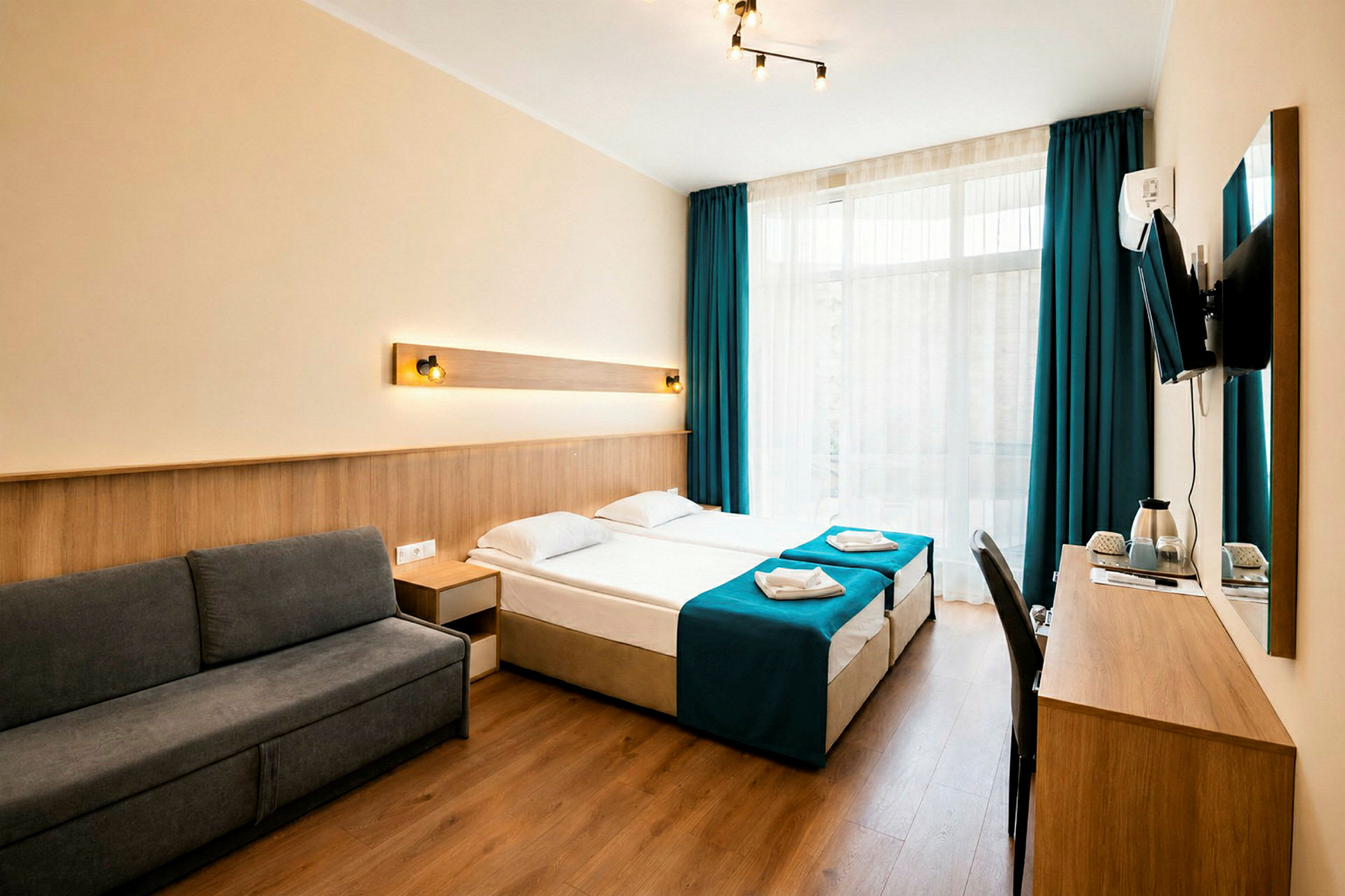 Et hotellrom med moderne innredning, mørkegrå sofa, to enkeltsenger med blå sengetepper og et skrivebord med en veggmontert TV. (AI-generert tekst)