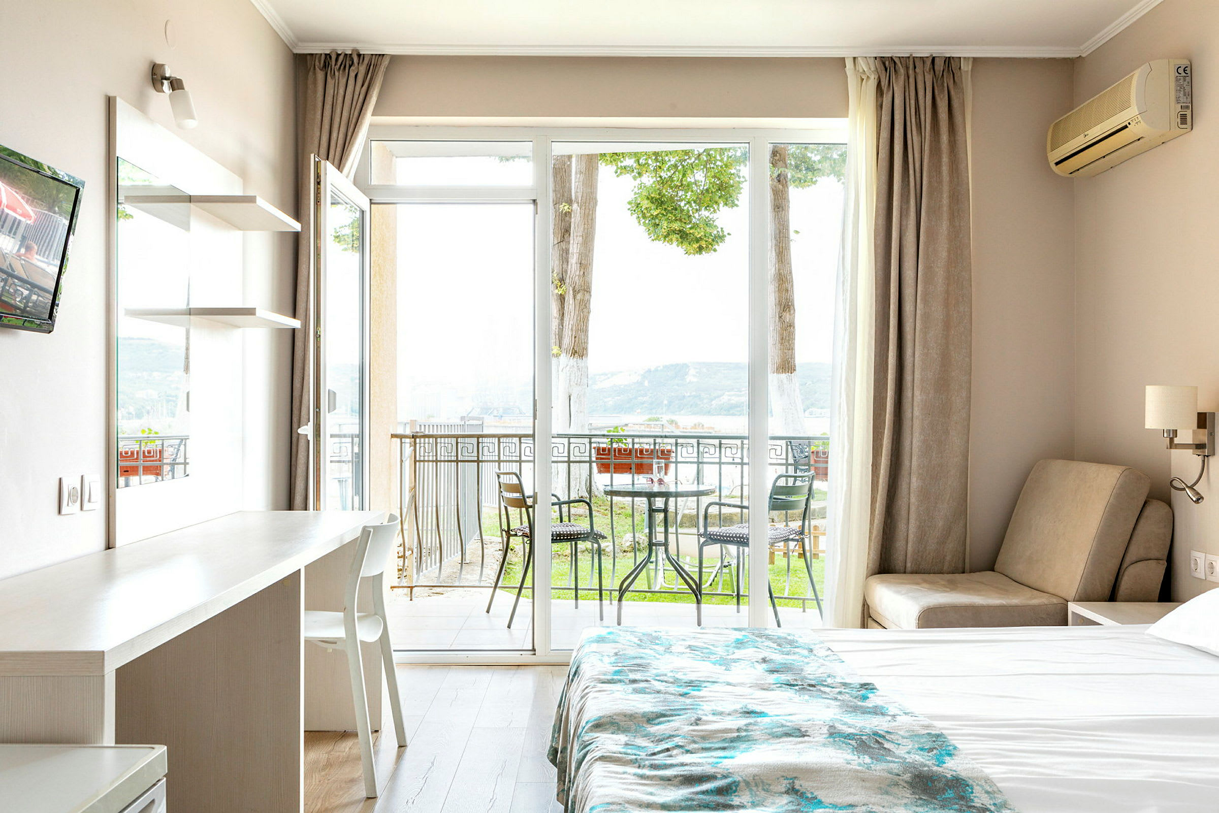 Et hotellrom med en seng, et bord og en stol, samt en balkong med utemøbler og utsikt mot naturen. (AI-generert tekst)