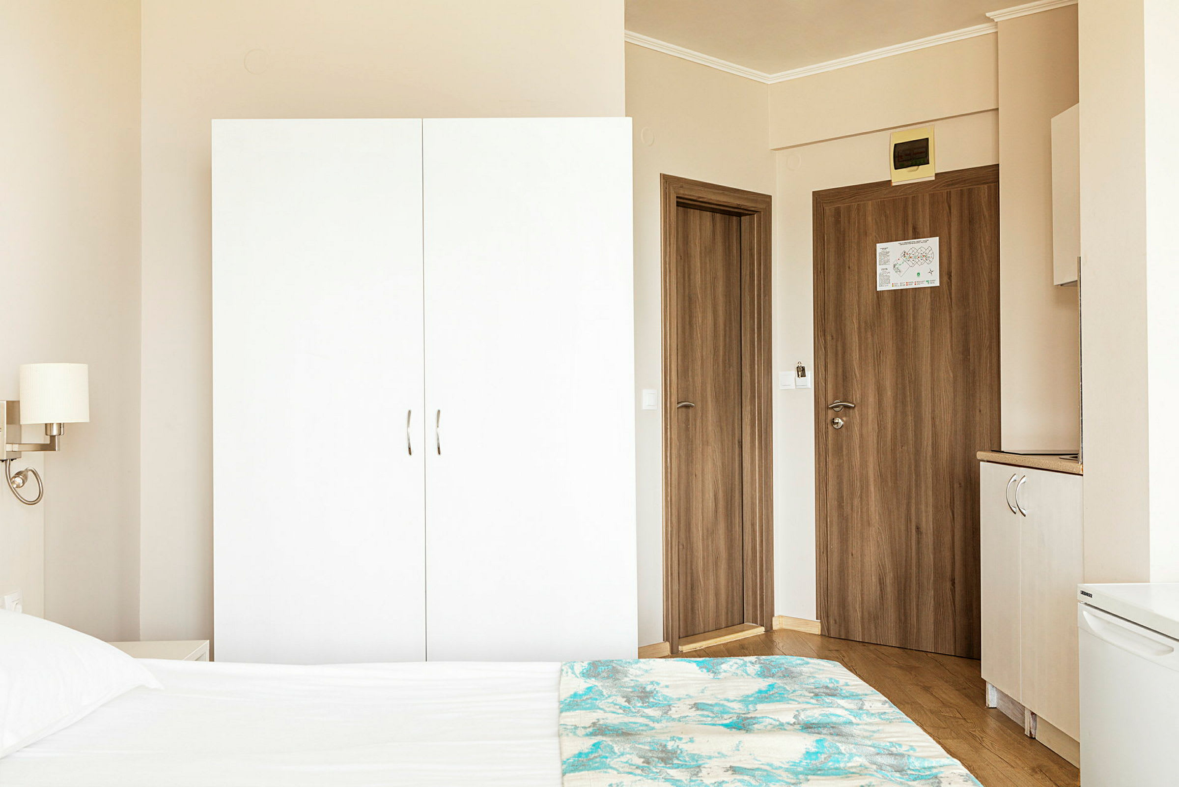 Et hotellromsmiljø med en hvit garderobe, tredører og en del av en seng i forgrunnen. (AI-generert tekst)