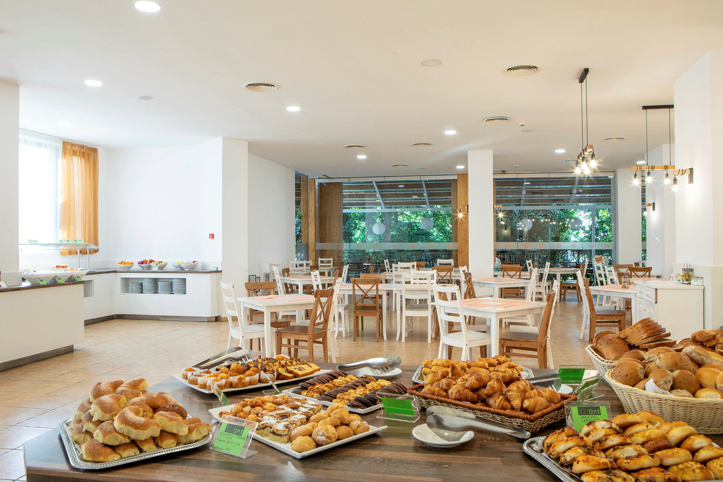 Et hotellrestaurantområde med bord og en buffet fylt med ulike brød og bakverk. (AI-generert tekst)
