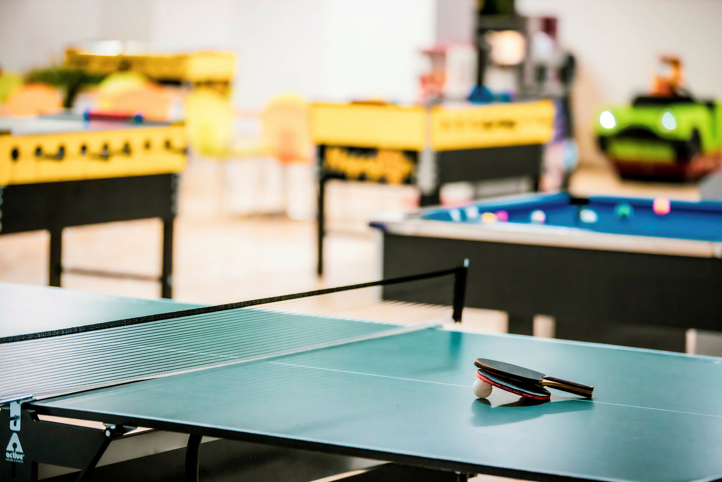 Et bordtennisbord med racketer og ball, med biljard- og bordfotballbord i bakgrunnen. (AI-generert tekst)