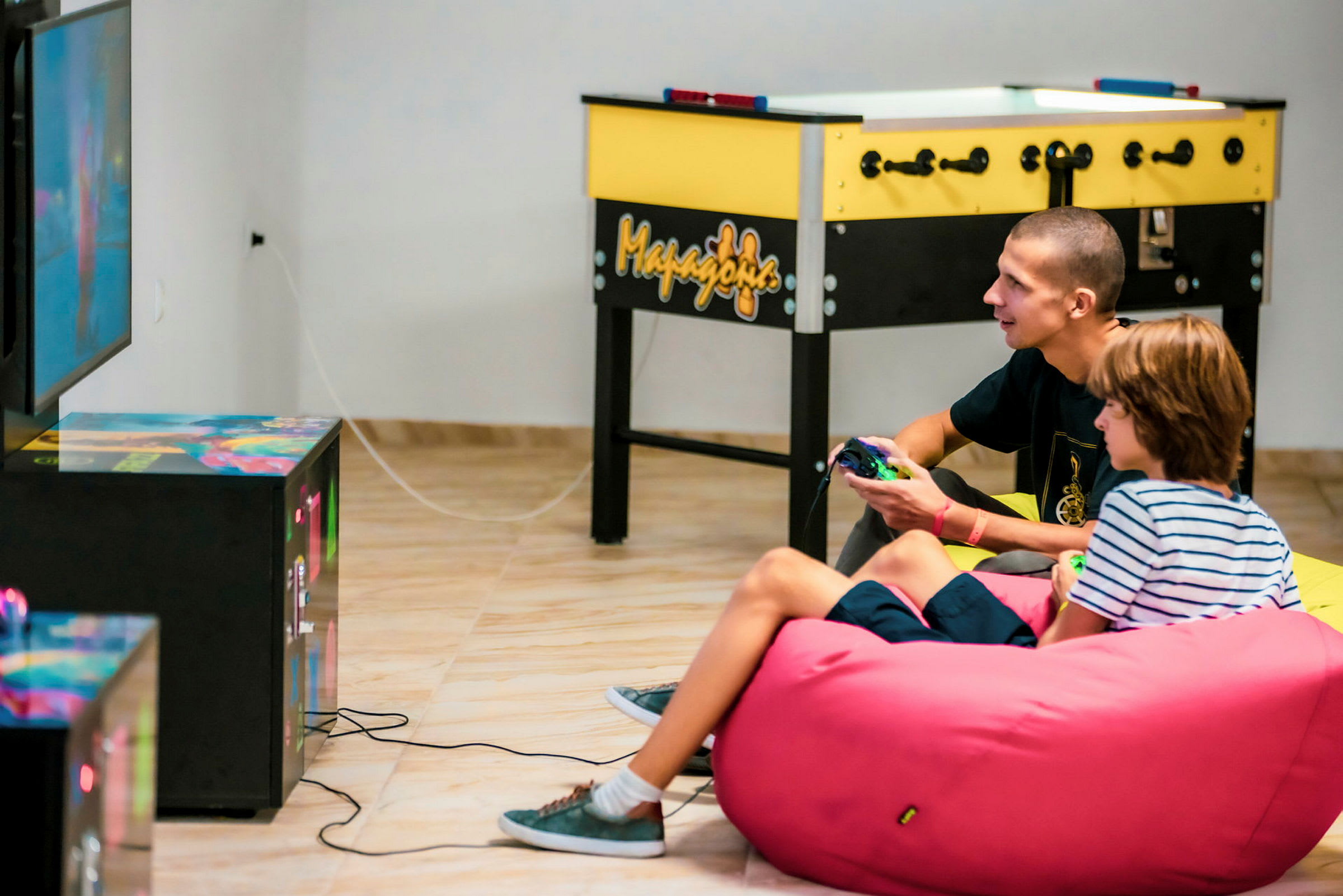 To personer spiller TV-spill og sitter på en rosa sittesekk. I bakgrunnen sees et bordfotballspill. (AI-generert tekst)