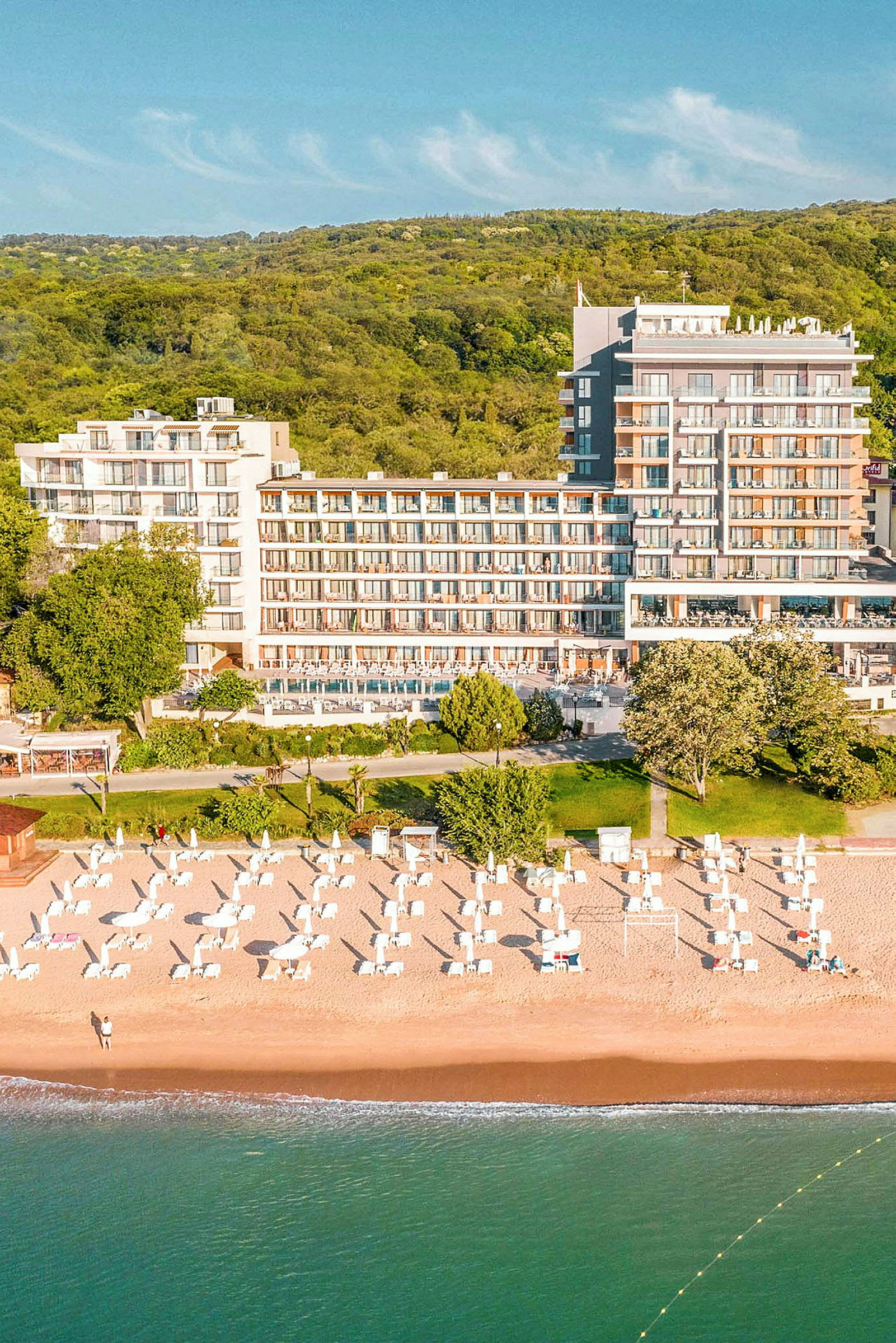 Utsikt over en strand med solsenger og parasoller ved et hotell, omgitt av grønne områder. (AI-generert tekst)