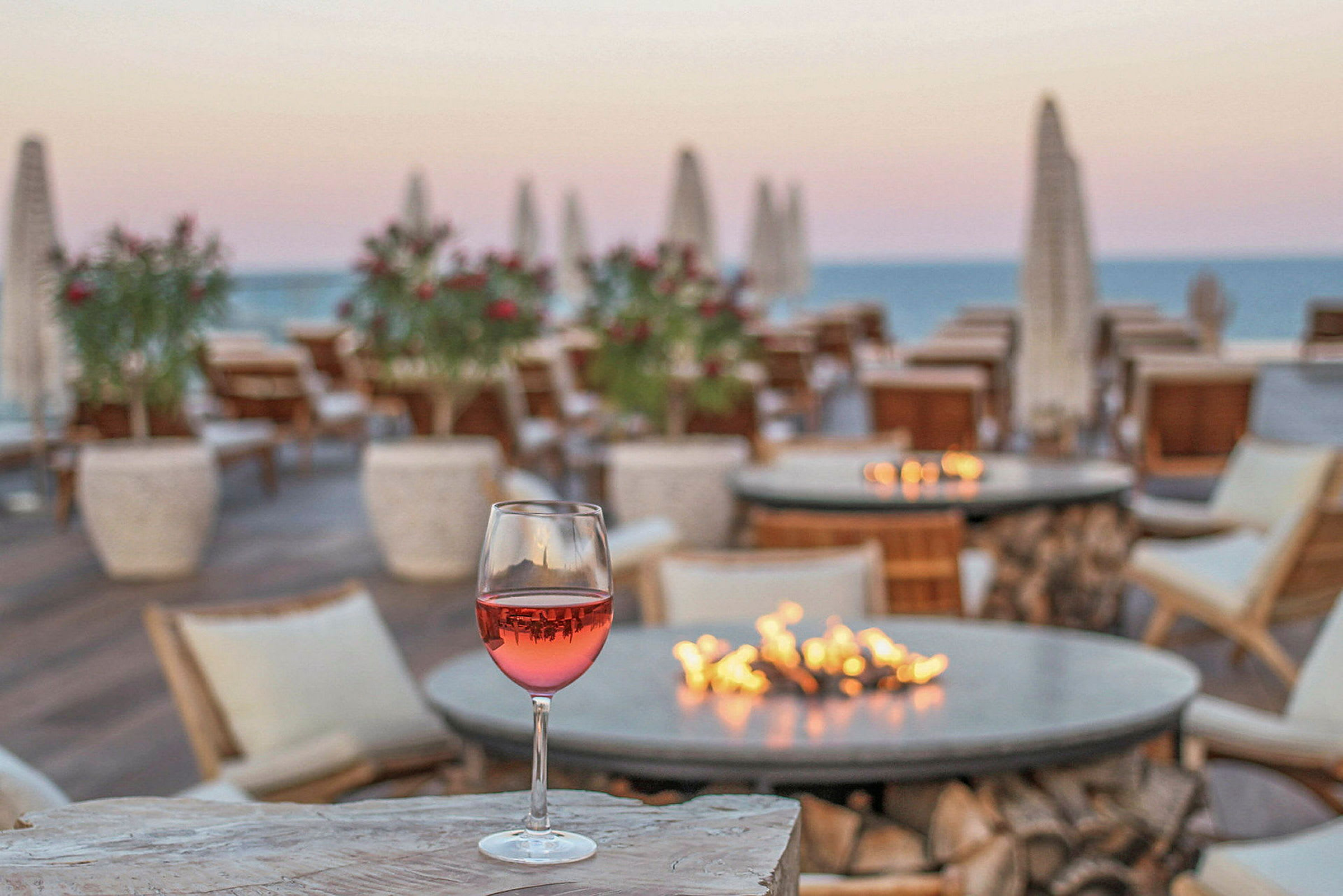 Et glass rosévin står på et steinbord i en utendørsrestaurant med utsikt mot sjøen og solnedgangen. (AI-generert tekst)