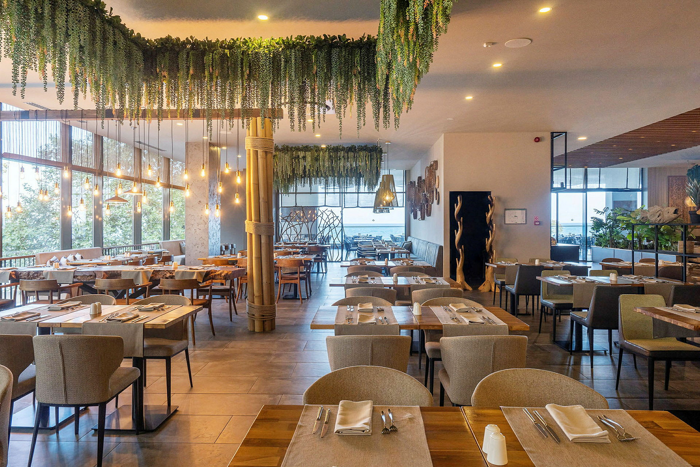En restaurant med bord dekket for servering, omgitt av grønn dekorasjon og lyskilder. (AI-generert tekst)