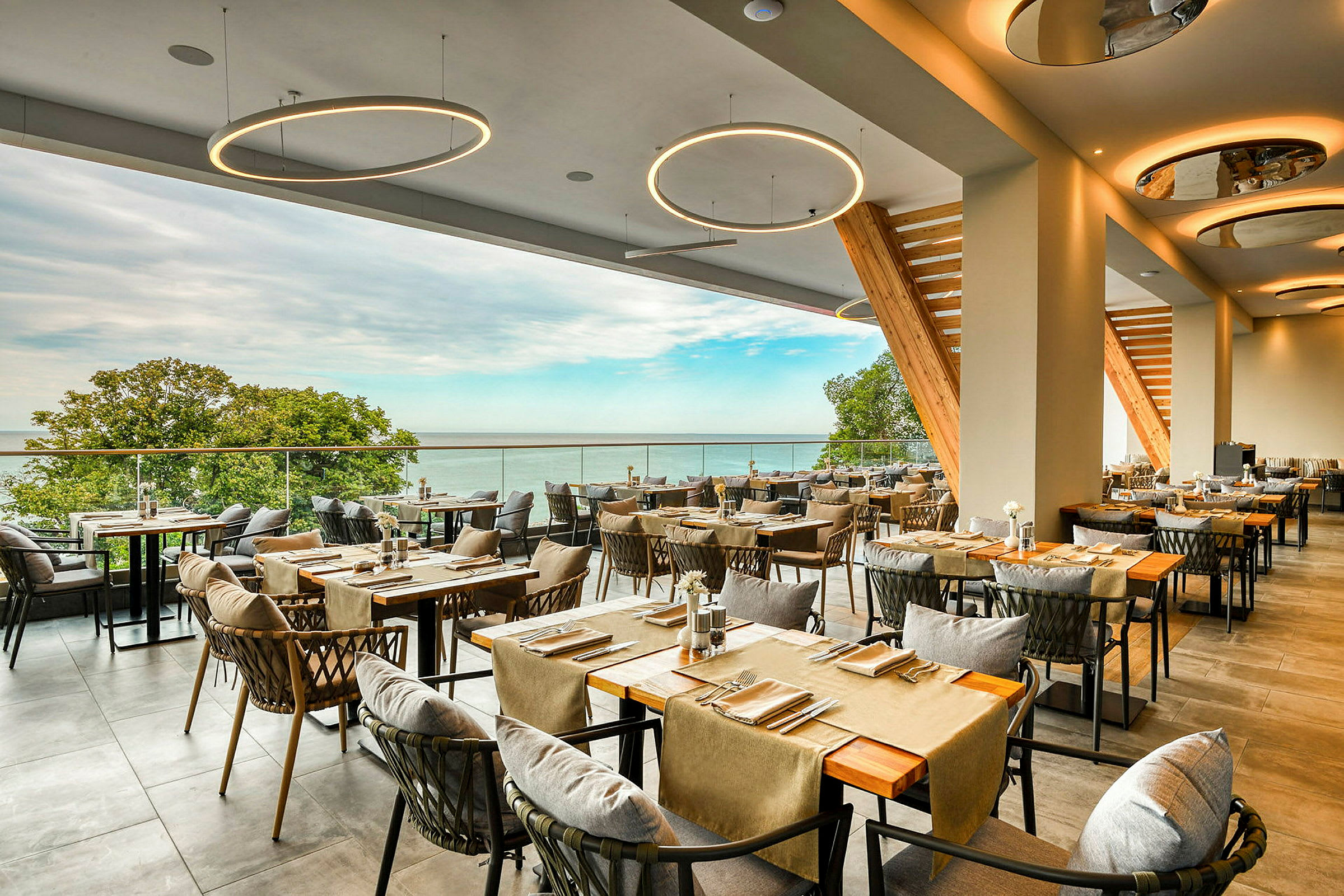 Restaurant med moderne innredede bord og stoler, samt utsikt mot havet og frodig natur. (AI-generert tekst)