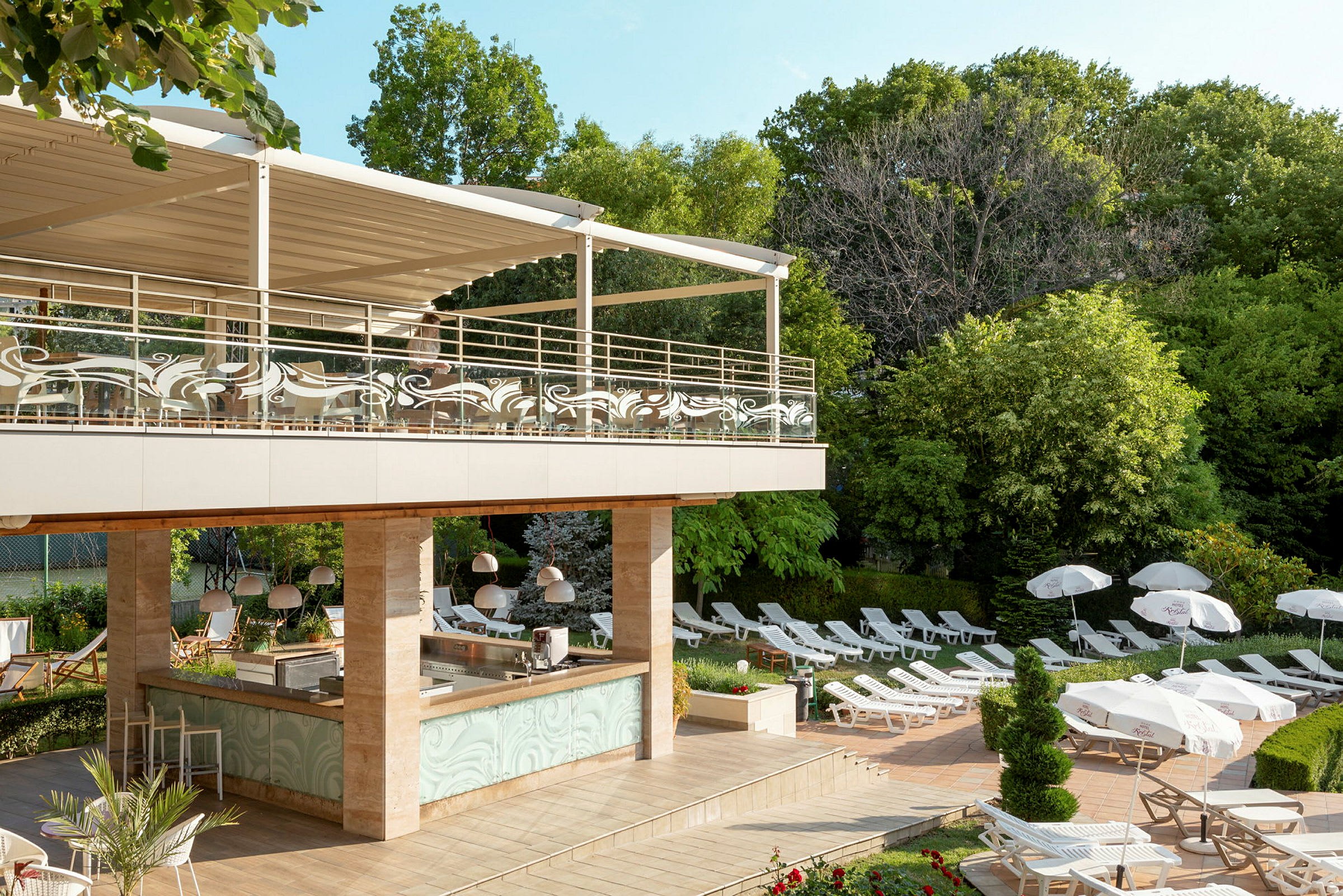 Et utendørs bassengområde med solsenger, parasoller og en bar under et overbygd terrasseområde. Området er omgitt av grønne trær. (AI-generert tekst)