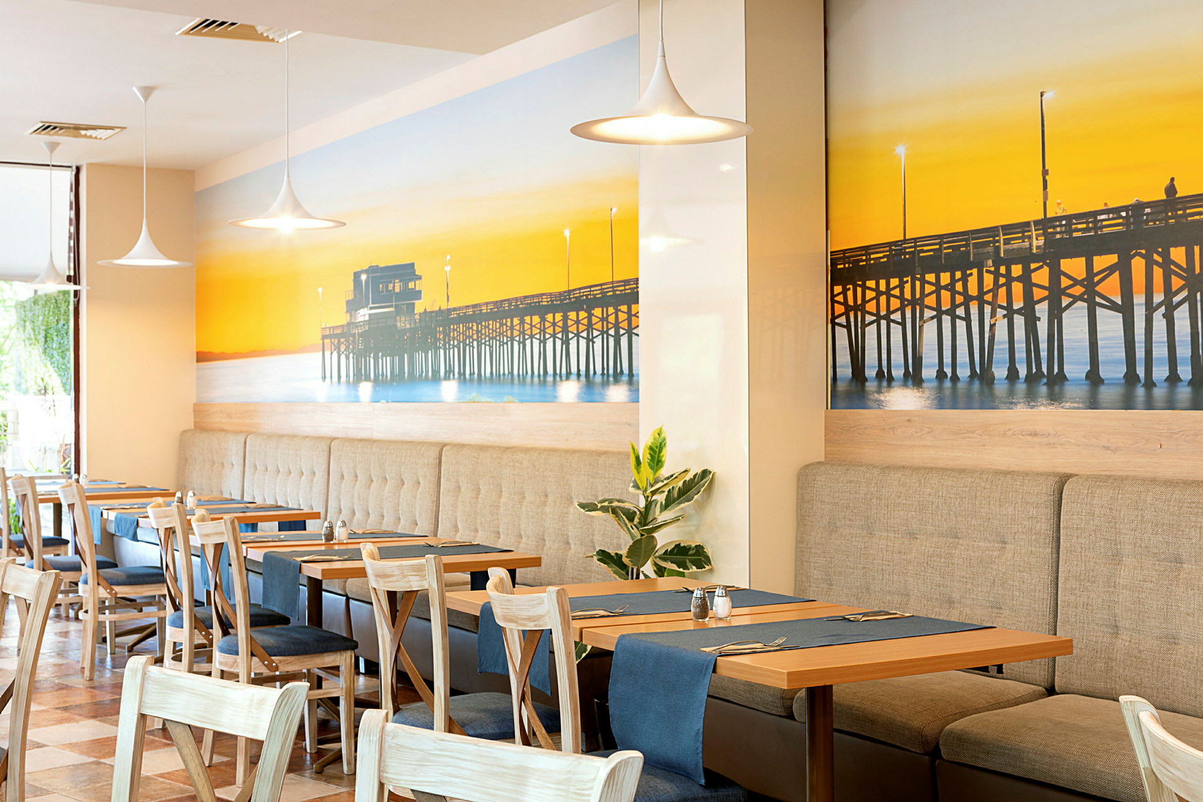 En restaurant med interiør som inkluderer bord og stoler, samt en veggdekorasjon som viser en brygge ved solnedgang. (AI-generert tekst)