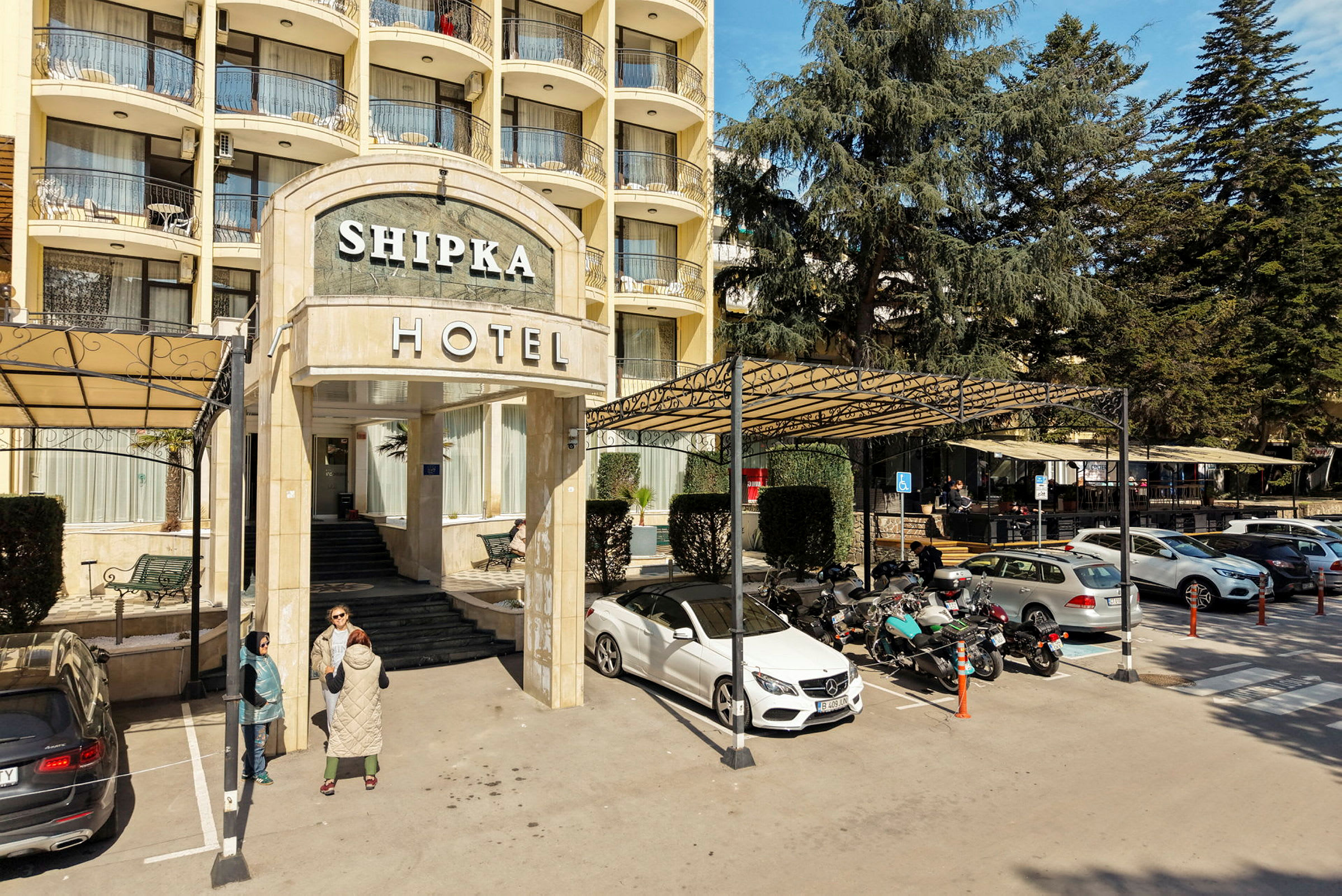 Inngangen til hotellbygningen med trapp og skilt, omgitt av parkering med biler og motorsykler. (AI-generert tekst)