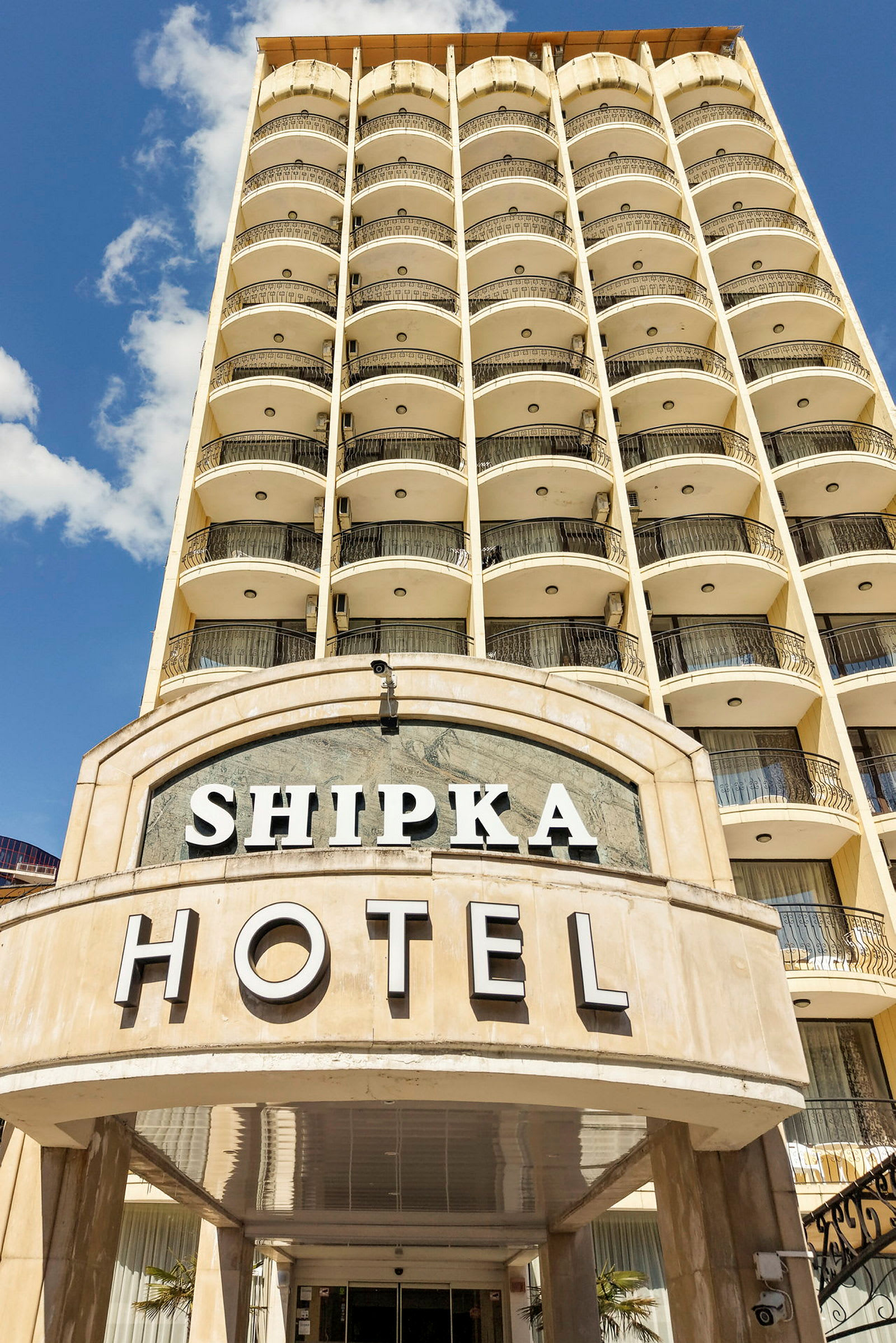 Hotellinngang med navnet Shipka, fleretasjes bygning i bakgrunnen. (AI-generert tekst)