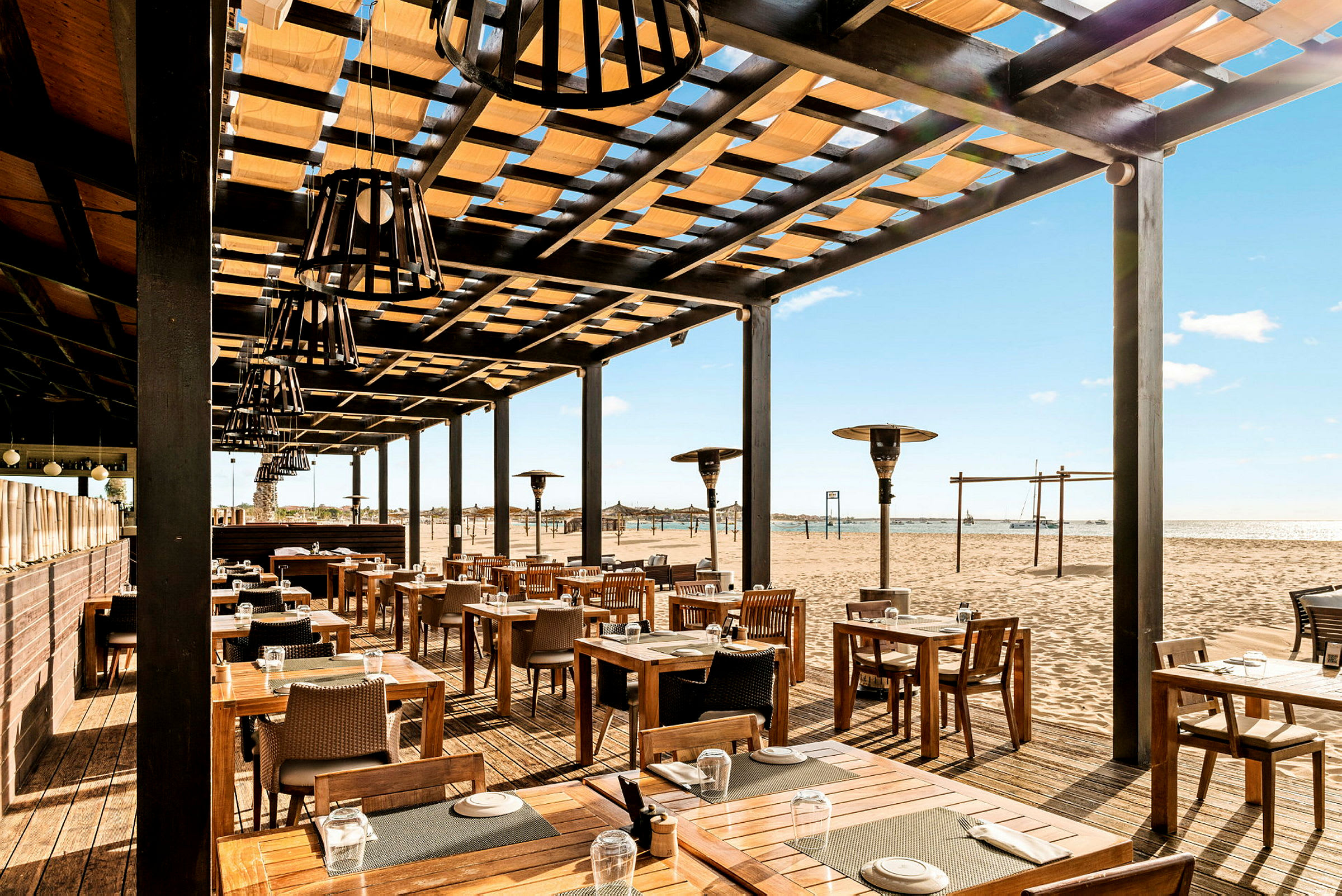 Utendørsrestaurant med trebord og stoler, plassert ved en sandstrand. (AI-generert tekst)