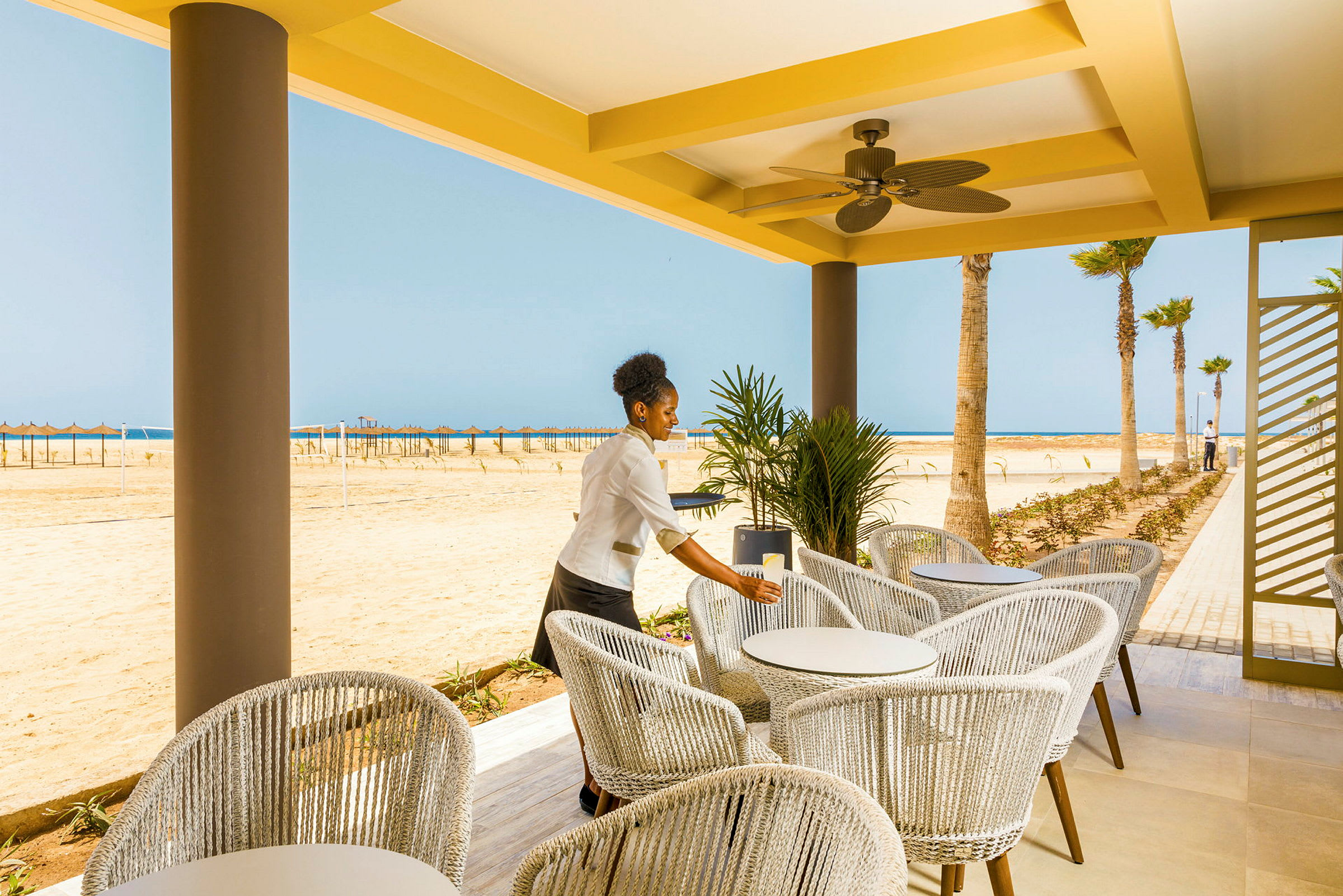 En uteterrasse med moderne møbler og utsikt over en sandstrand og havet i bakgrunnen. (AI-generert tekst)