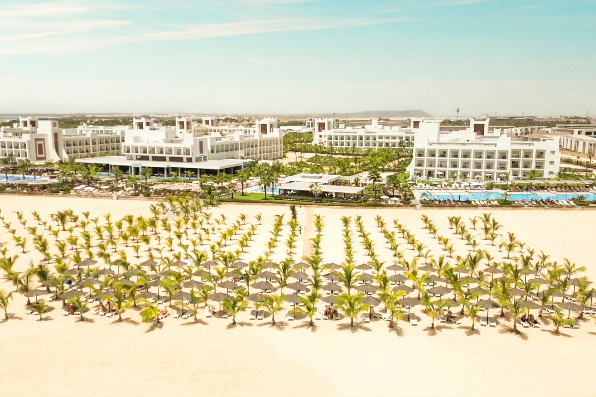 Sandstrand med parasoller og solsenger, omgitt av hvite hotellbygg og grøntområder. (AI-generert tekst)