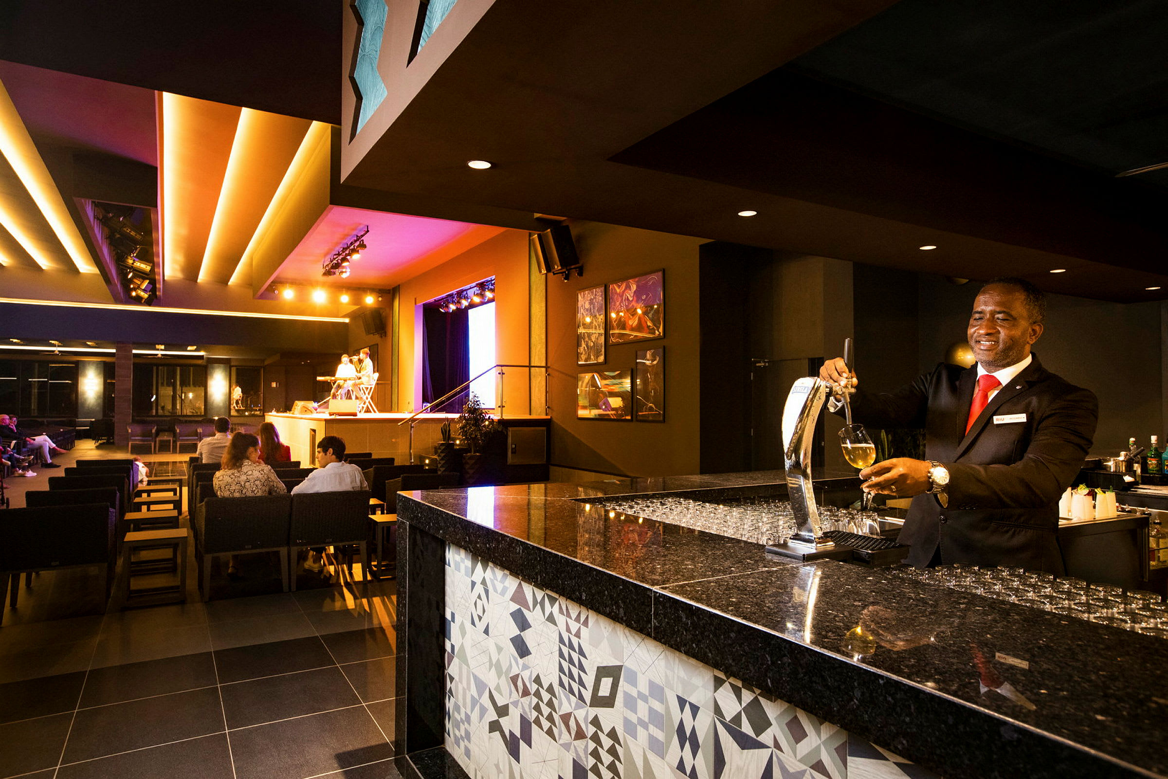 En elegant bar med en bartender som serverer drikke og en opplyst scene i bakgrunnen med sittende gjester. (AI-generert tekst)