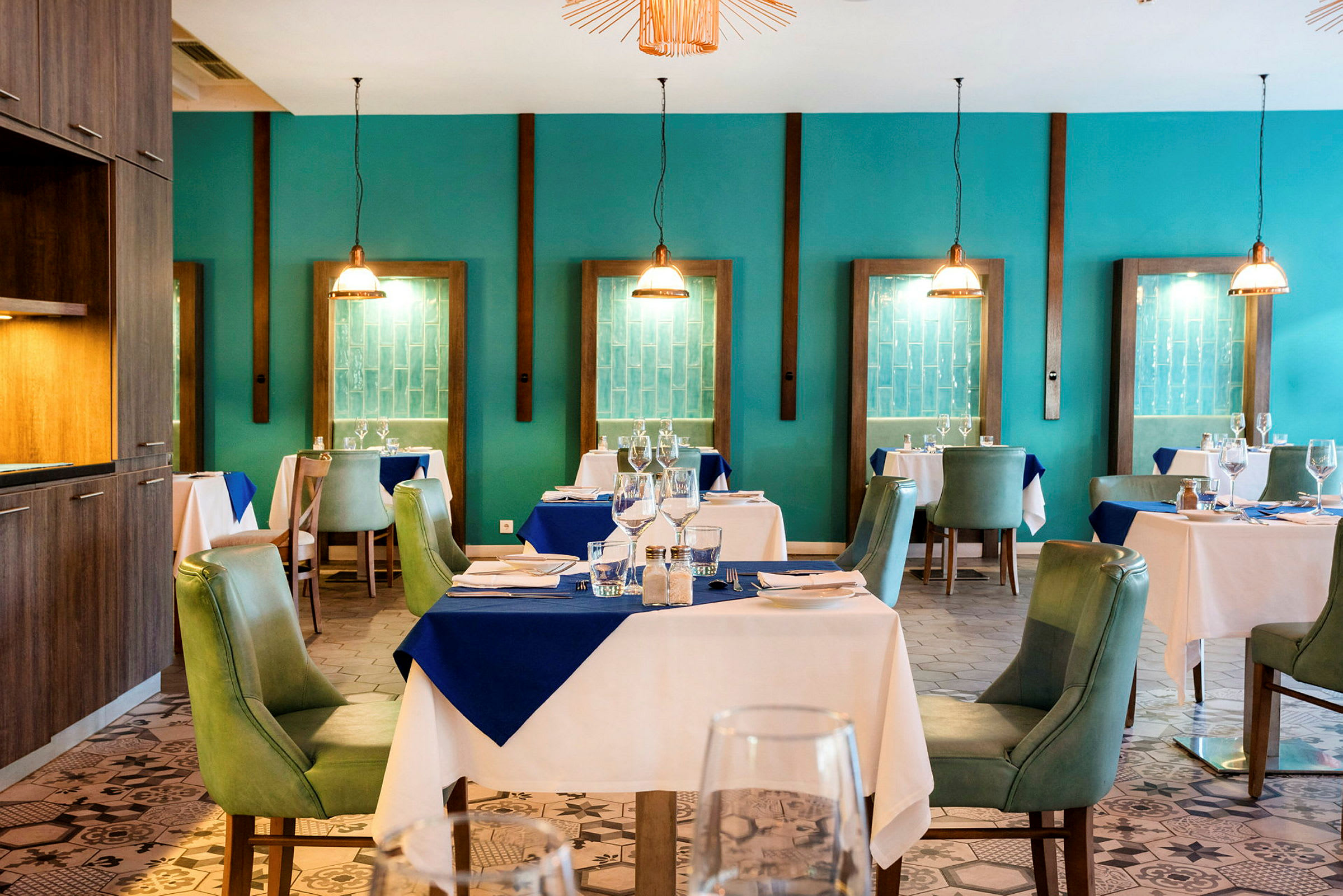 Et elegant restaurantinteriør med dekket bord og grønne stoler, omgitt av turkise vegger og dekorative hengelamper. (AI-generert tekst)