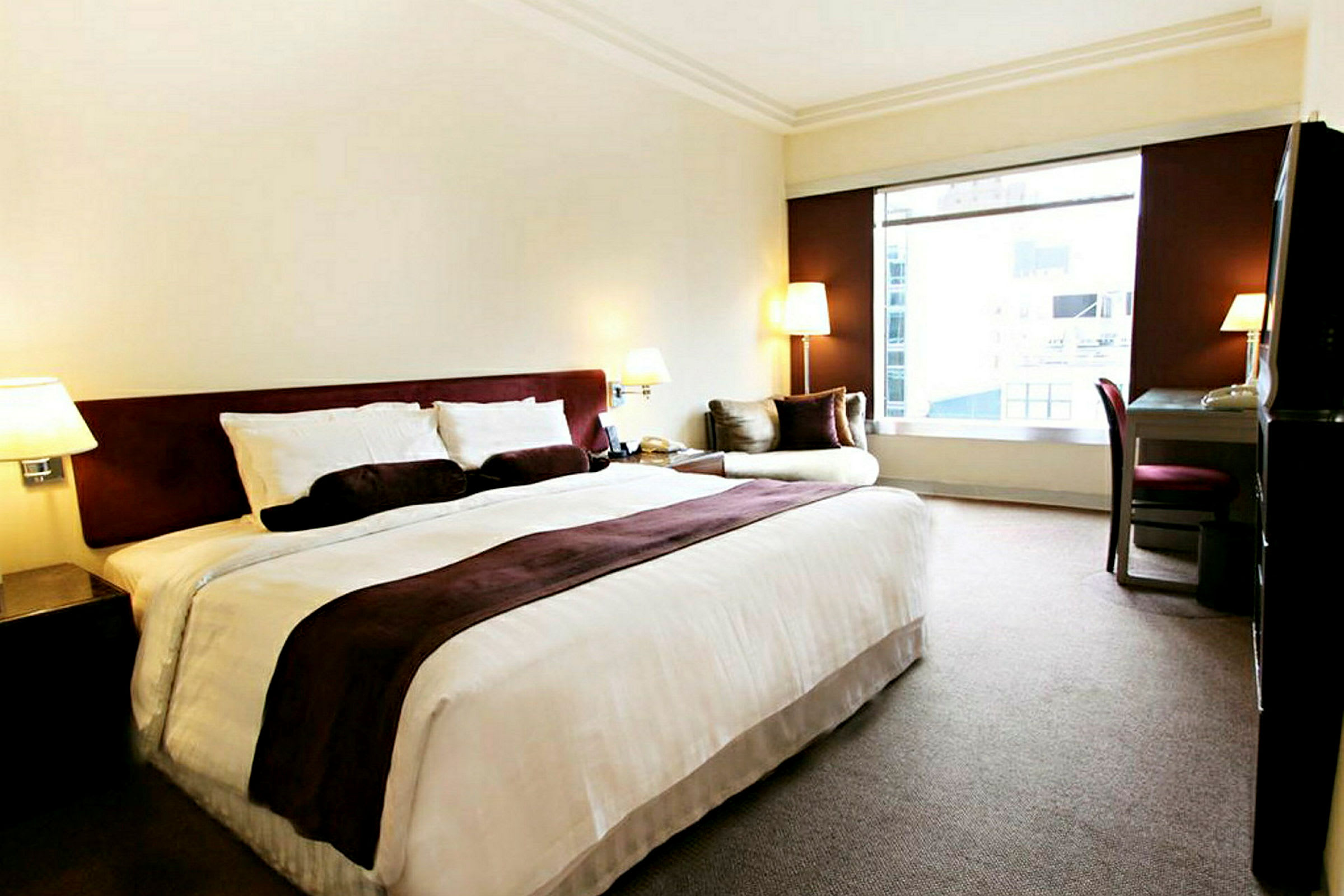 Hotellrom med en stor seng, et skrivebord og en lenestol ved siden av et stort vindu. (AI-generert tekst)