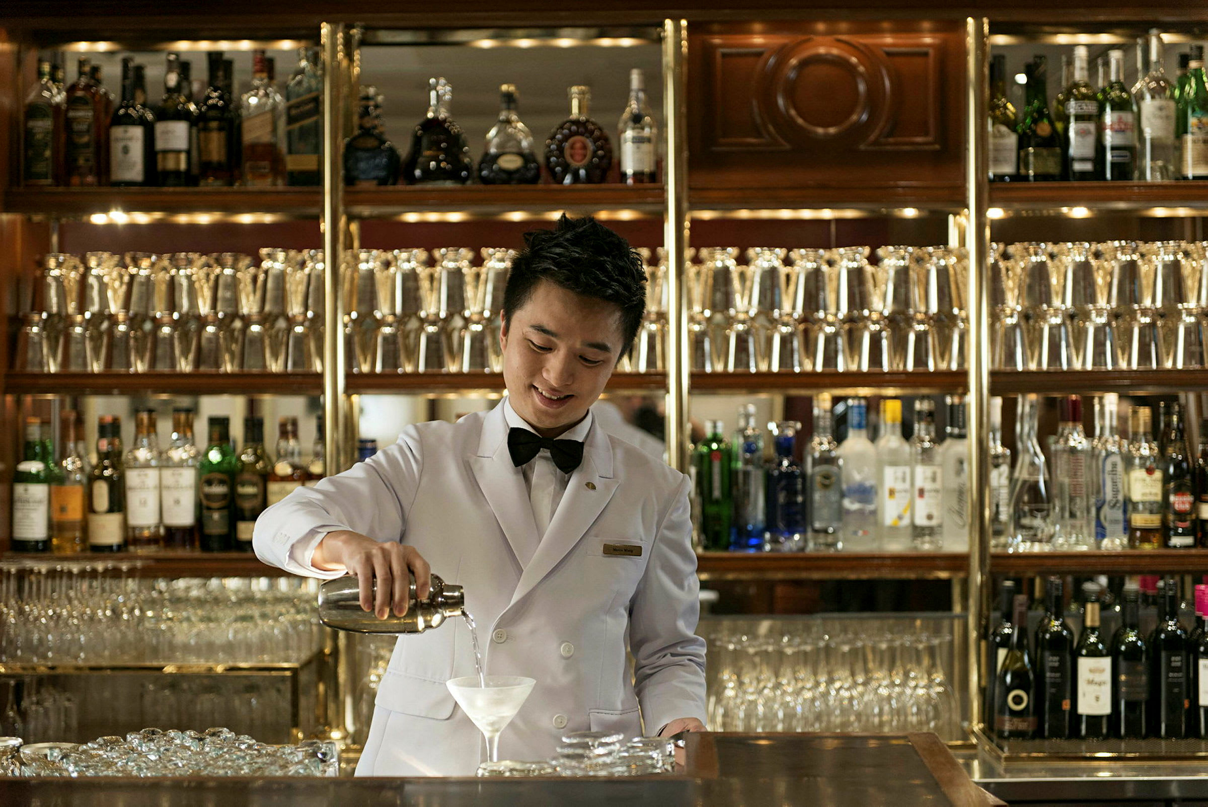 En bartender kledd i hvit jakke som serverer en drink foran en velfylt bar. (AI-generert tekst)