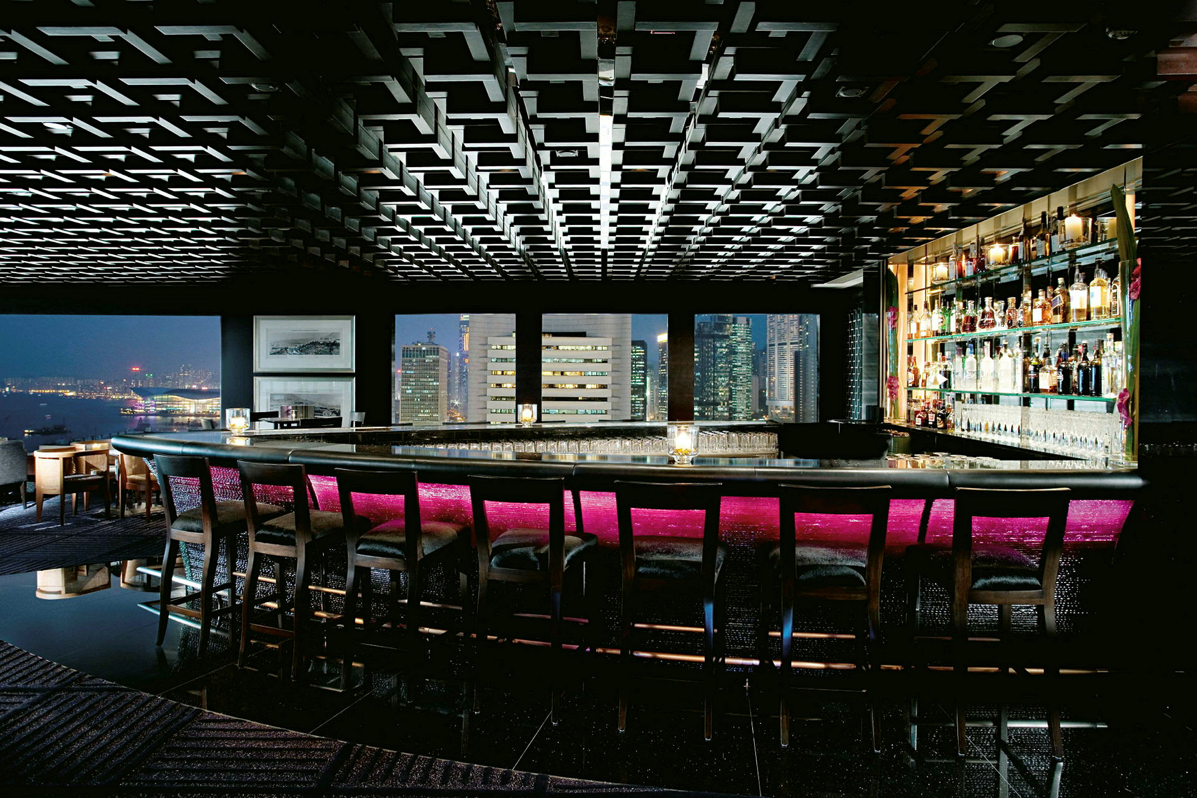 Bilde av en luksuriøs bar med høye ryggstoler ved bardisken. Baren har stemningsfull belysning med en neonrosa nyanse, med store vinduer som viser utsikten over byens skyline. (AI-generert tekst)