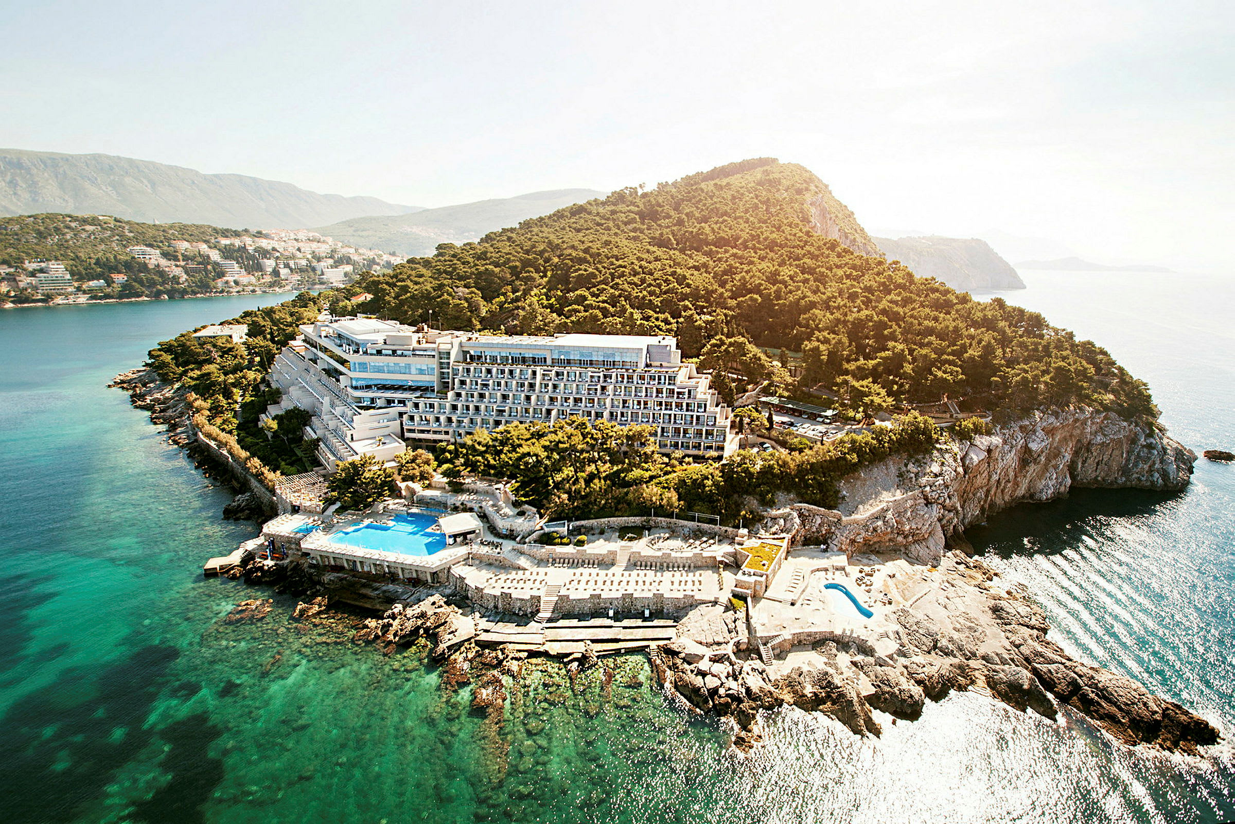 Flyfoto av en hotellbygning beliggende ved en klippefylt kyst omgitt av trær og hav. (AI-generert tekst)