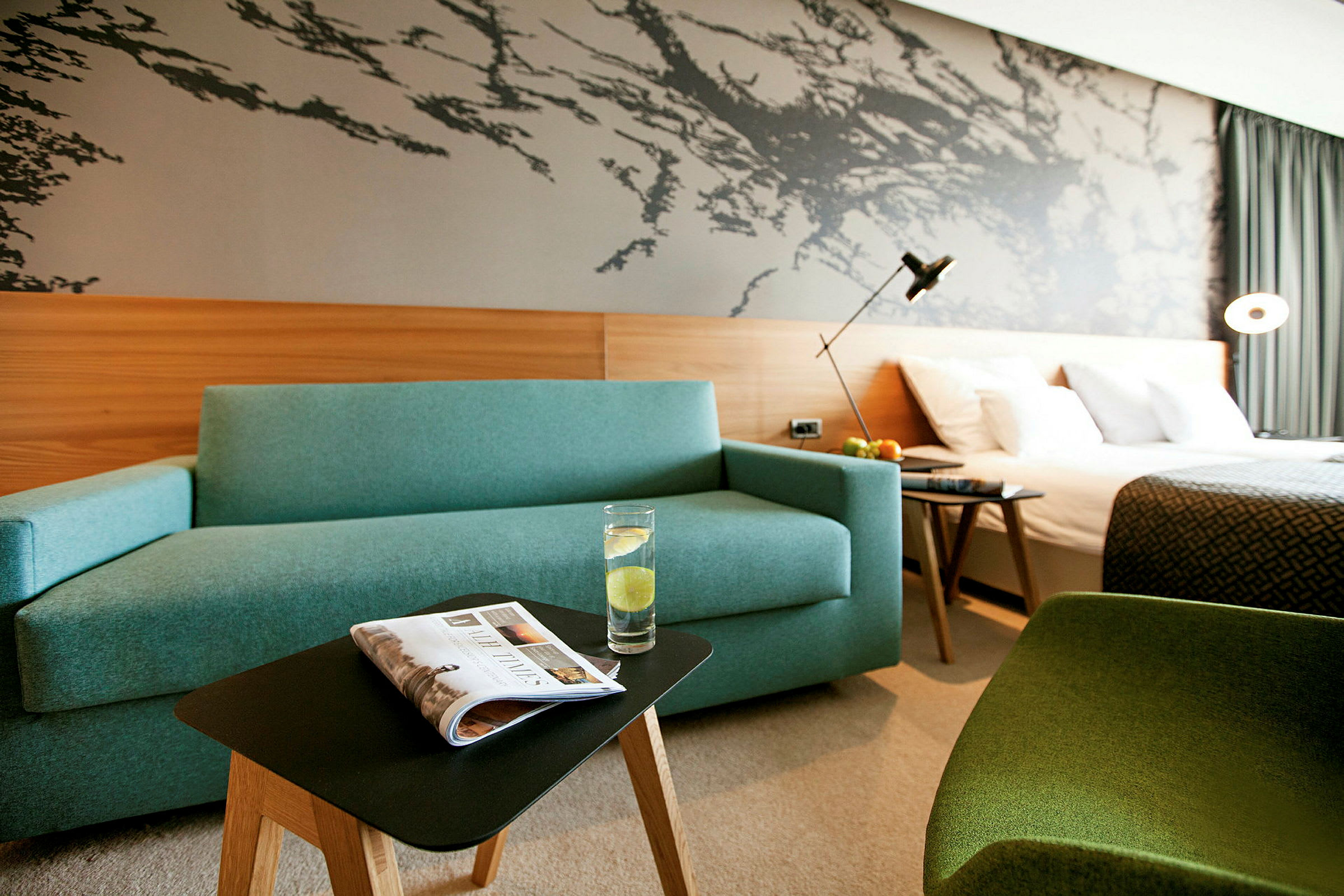 Hotellrom med en blå sofa, et lite bord med et glass vann, en seng og en dekorativ vegg. (AI-generert tekst)
