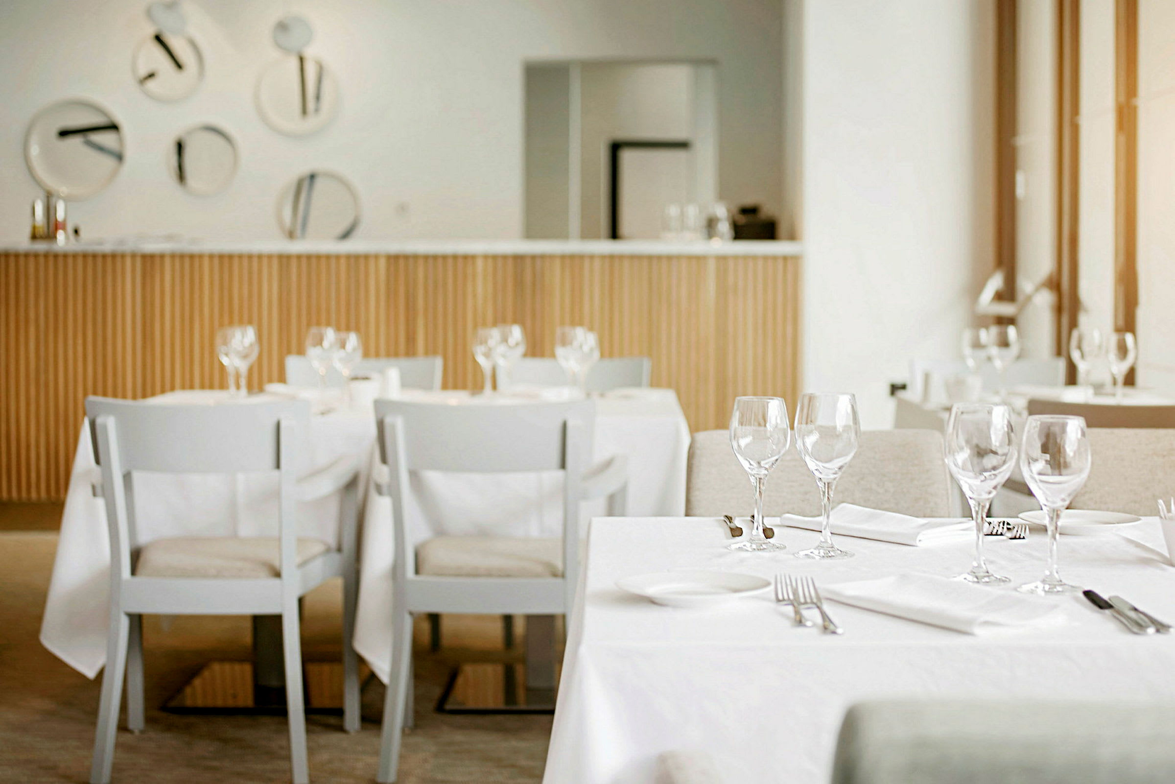 Et moderne og lyst restaurantinteriør med dekkede bord og hvite stoler. På bordene er glass, bestikk og servietter. (AI-generert tekst)