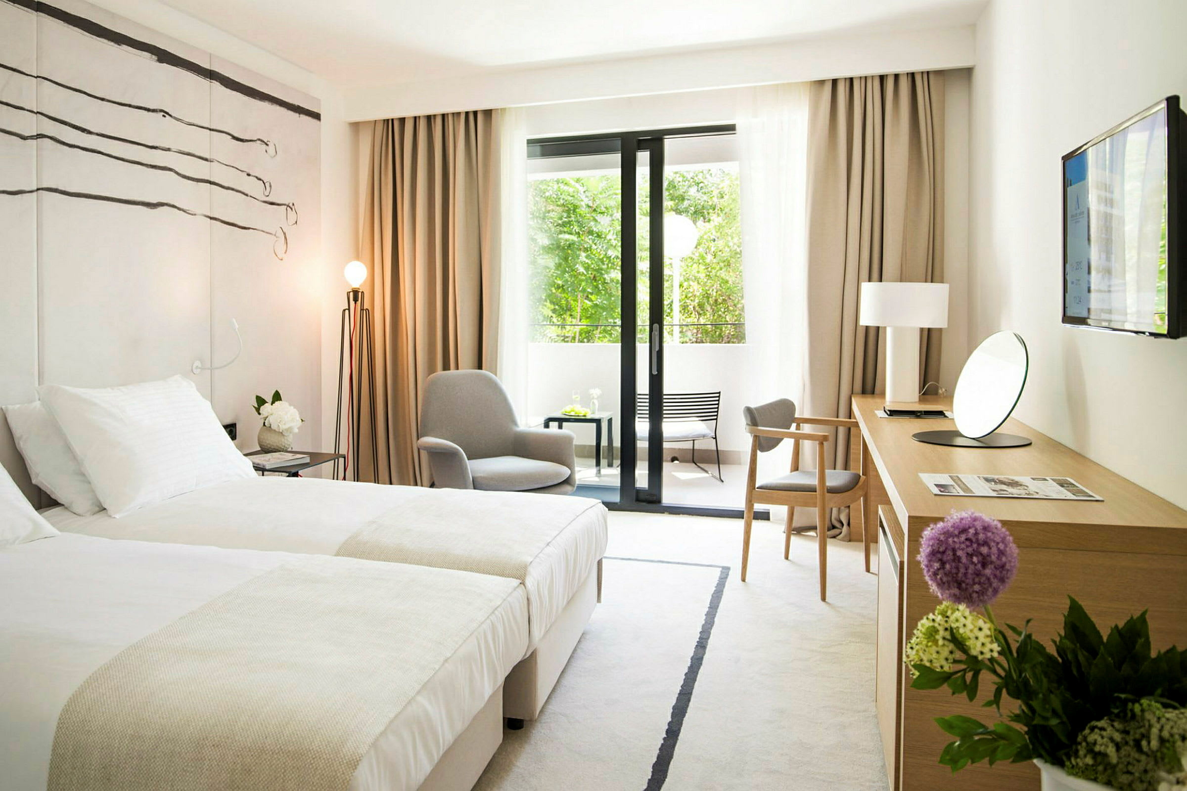 Et moderne hotellrom med to senger, en lenestol, et skrivebord med et speil og en TV på veggen. Det er store vindusdører som fører til en utendørs balkong med utsikt mot grøntområder. (AI-generert tekst)