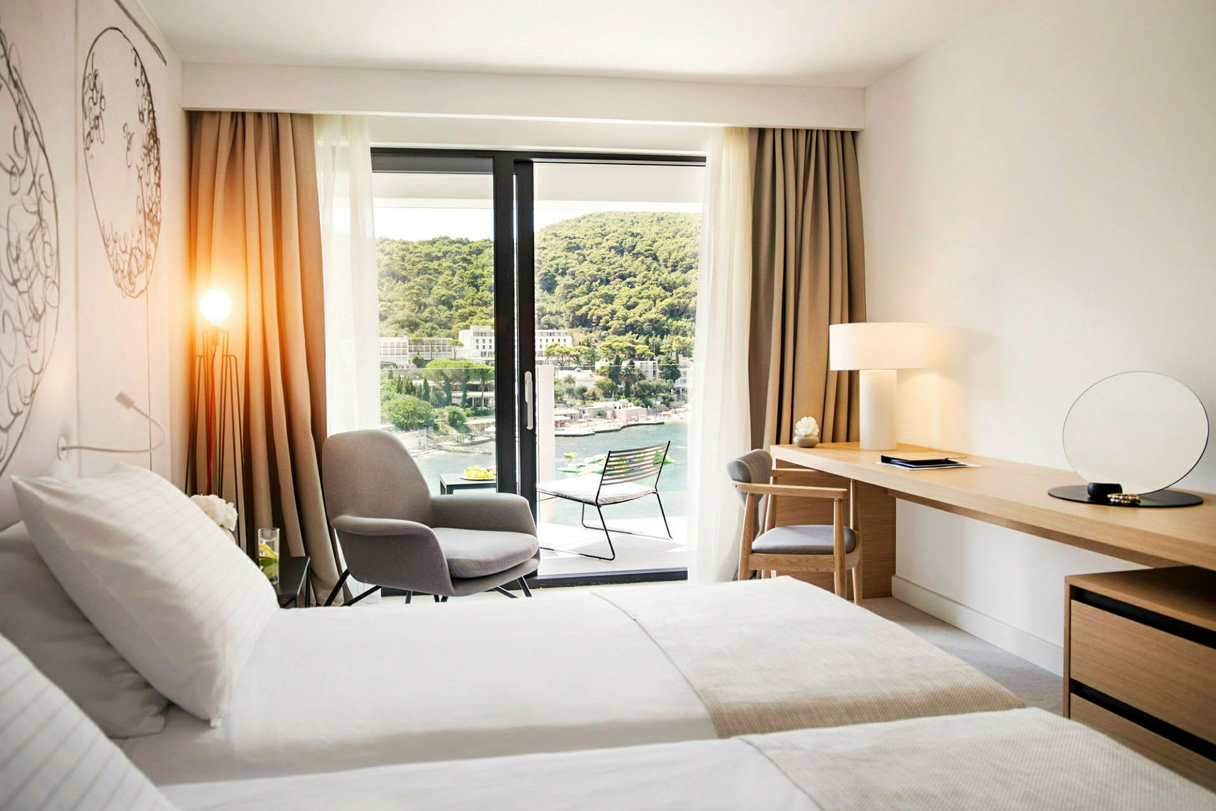 Moderne hotellrom med dobbeltseng, sitteområde, skrivebord og balkong med utsikt over en frodig åsside og vann. (AI-generert tekst)
