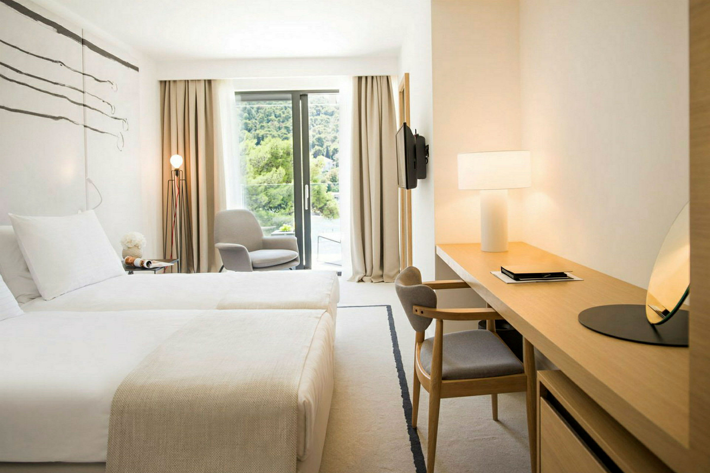 Et lyst hotellrom med to senger, et skrivebordshjørne, en stol ved et vindu og utsikt over grøntområder. (AI-generert tekst)