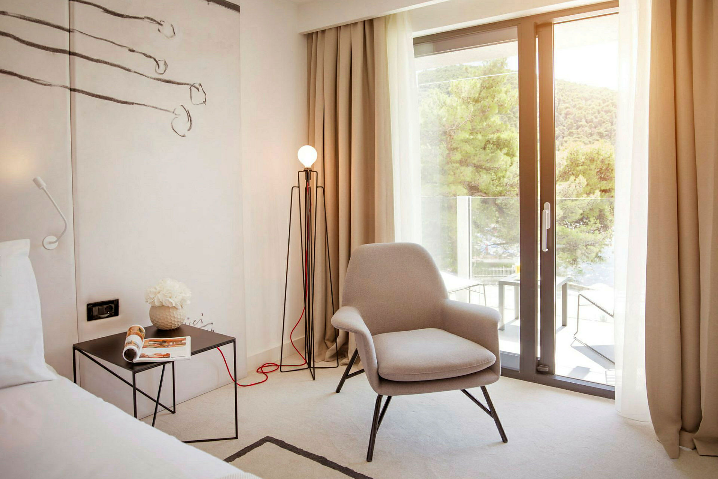 Et hotellrom med en komfortabel lenestol, et sidebord med et magasin og en gulvlampe. En stor glassdør åpner ut til en balkong med utsikt over frodige trær. (AI-generert tekst)