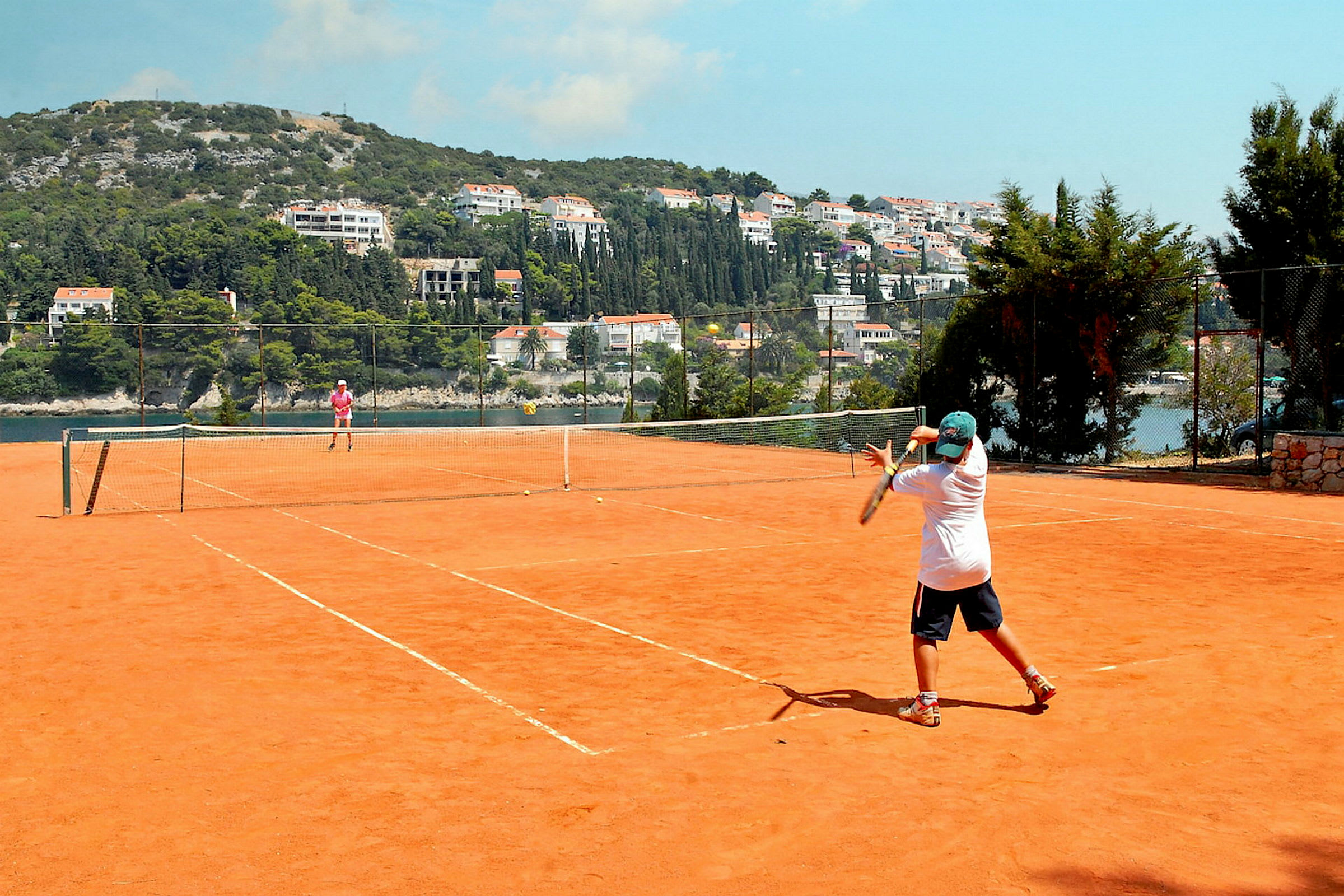 To personer spiller tennis på en utendørs grusbane. Bak tennisbanen kan man se en nærliggende ås med bygninger og grøntområder. (AI-generert tekst)