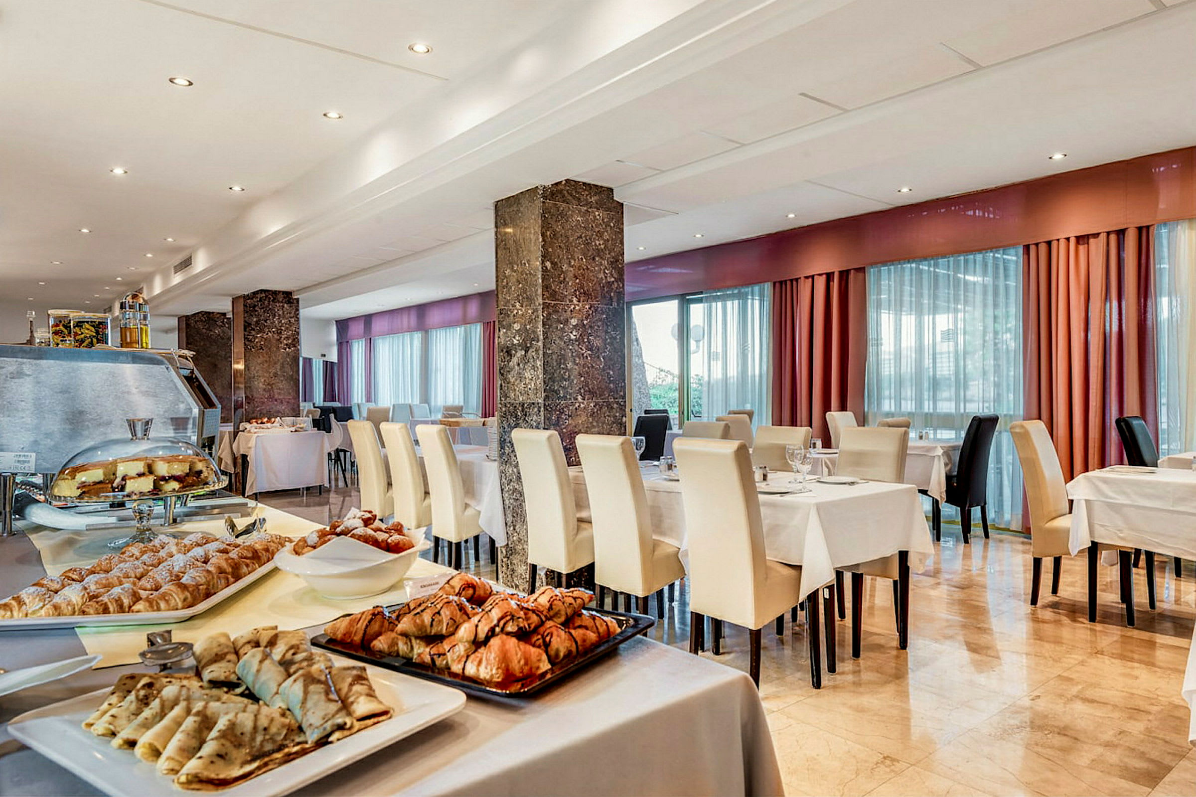 En hotellrestaurant med dekkede bord og hvite stoler, samt et frokostbuffébord med forskjellige bakverk og pannekaker. (AI-generert tekst)