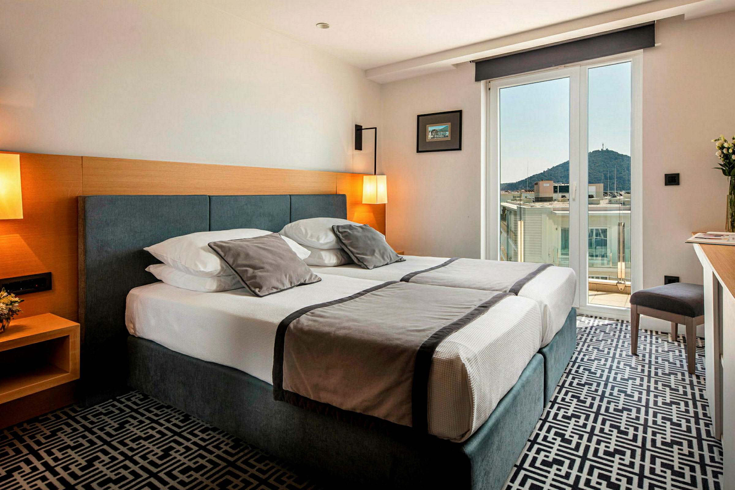 Hotellrom med dobbeltseng, lys innredning og balkong med utsikt over by og fjell. (AI-generert tekst)