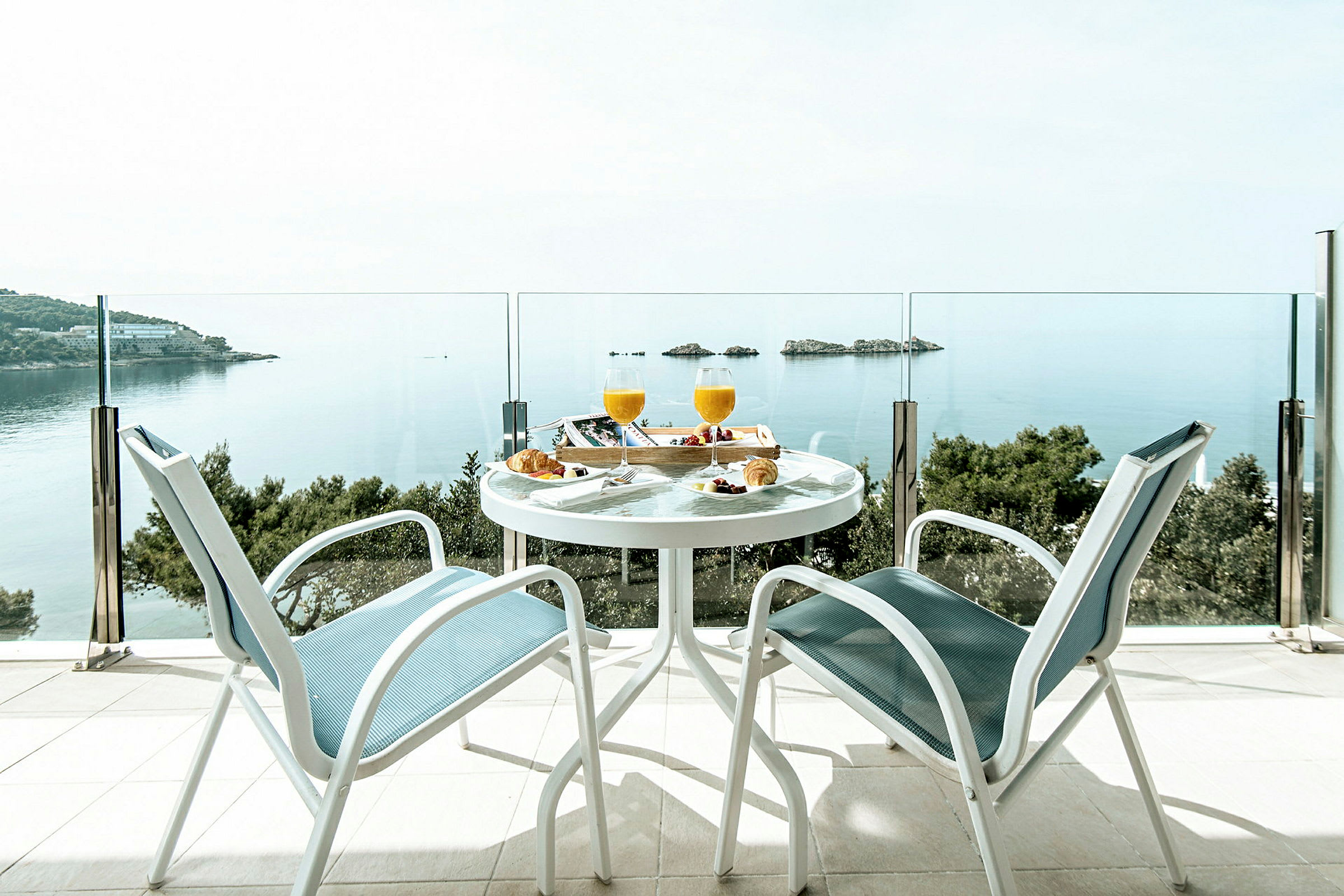 To stoler og et bord med frokost er plassert på en terrasse med utsikt over havet og noen små øyer. (AI-generert tekst)