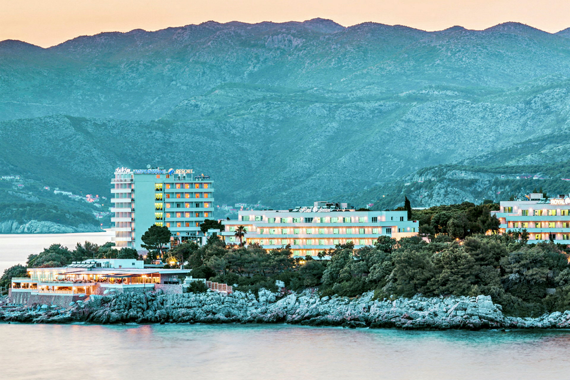 Et kystnært hotell med utsikt over havet, omgitt av trær og grønne områder, med fjell i bakgrunnen. (AI-generert tekst)